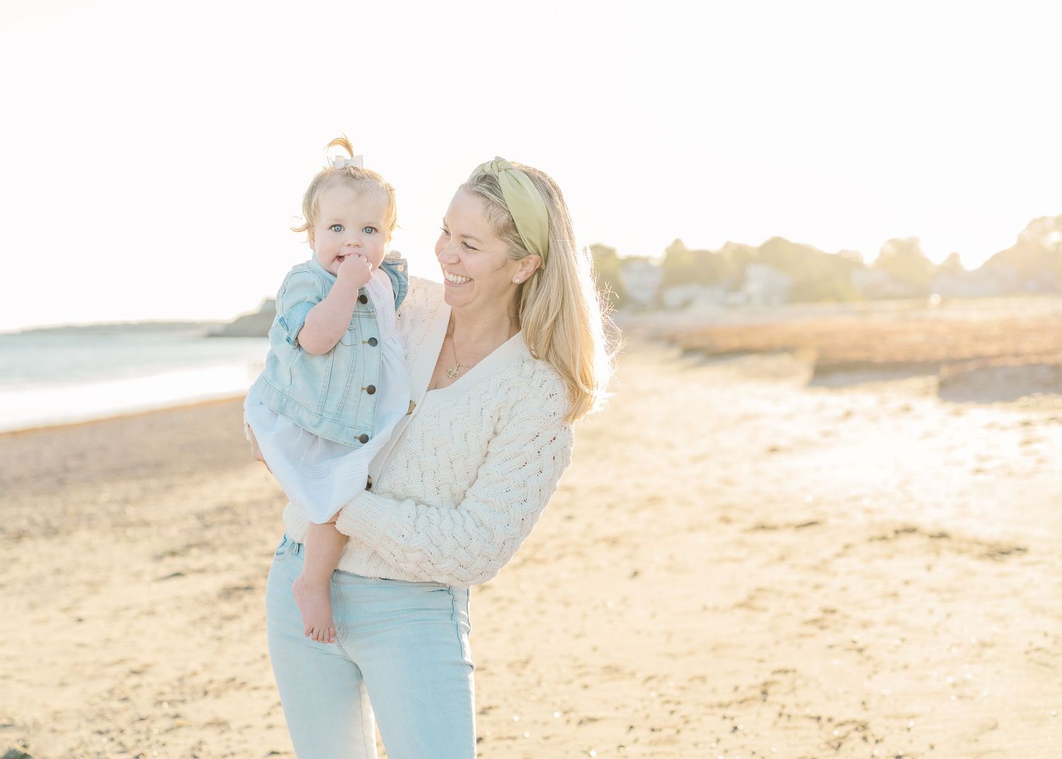 Mother in white sweater holding a small child on a sunny beach.