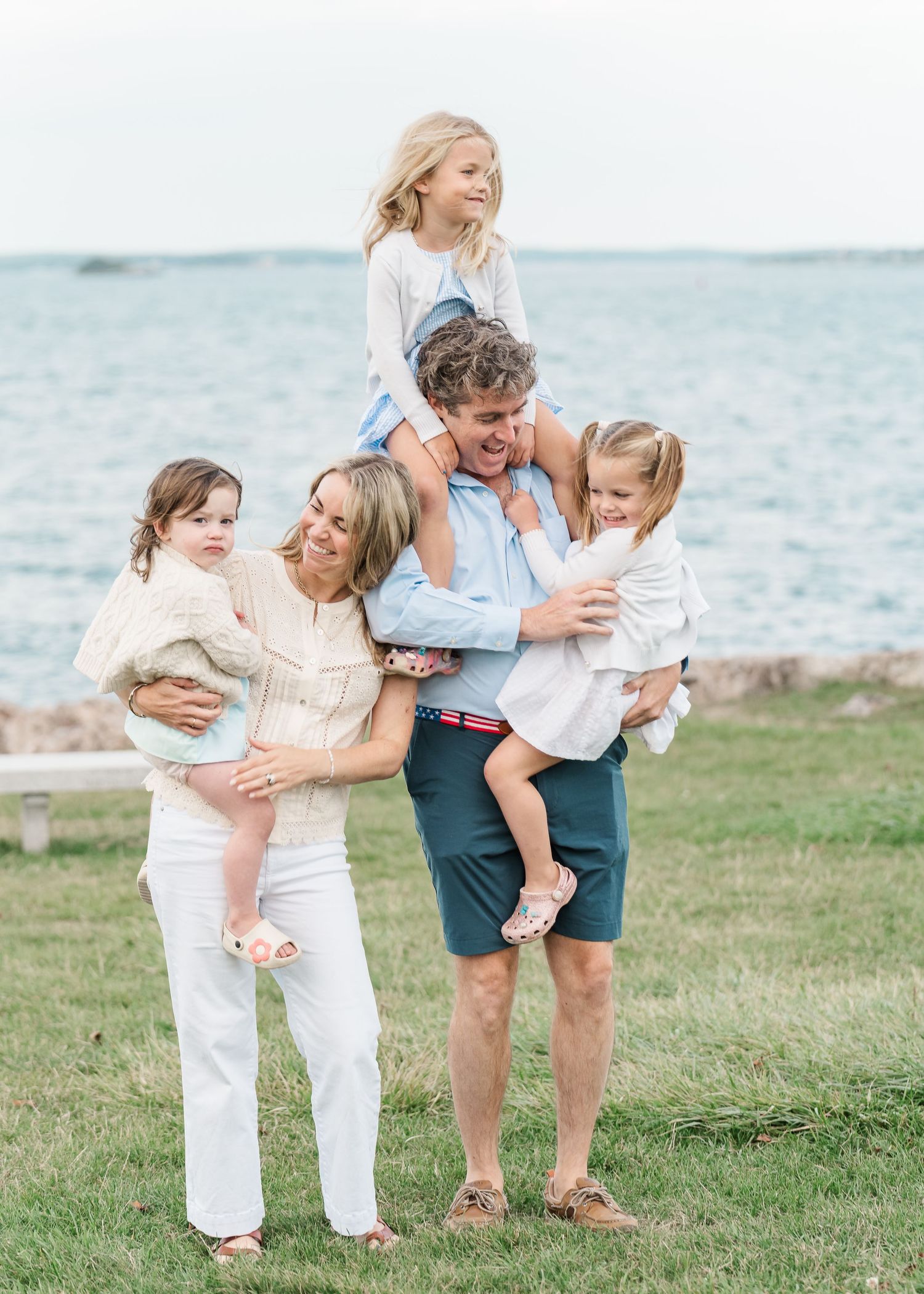 A family shares joyful embraces while posing together on a grassy oceanfront setting in coordinated white outfits.