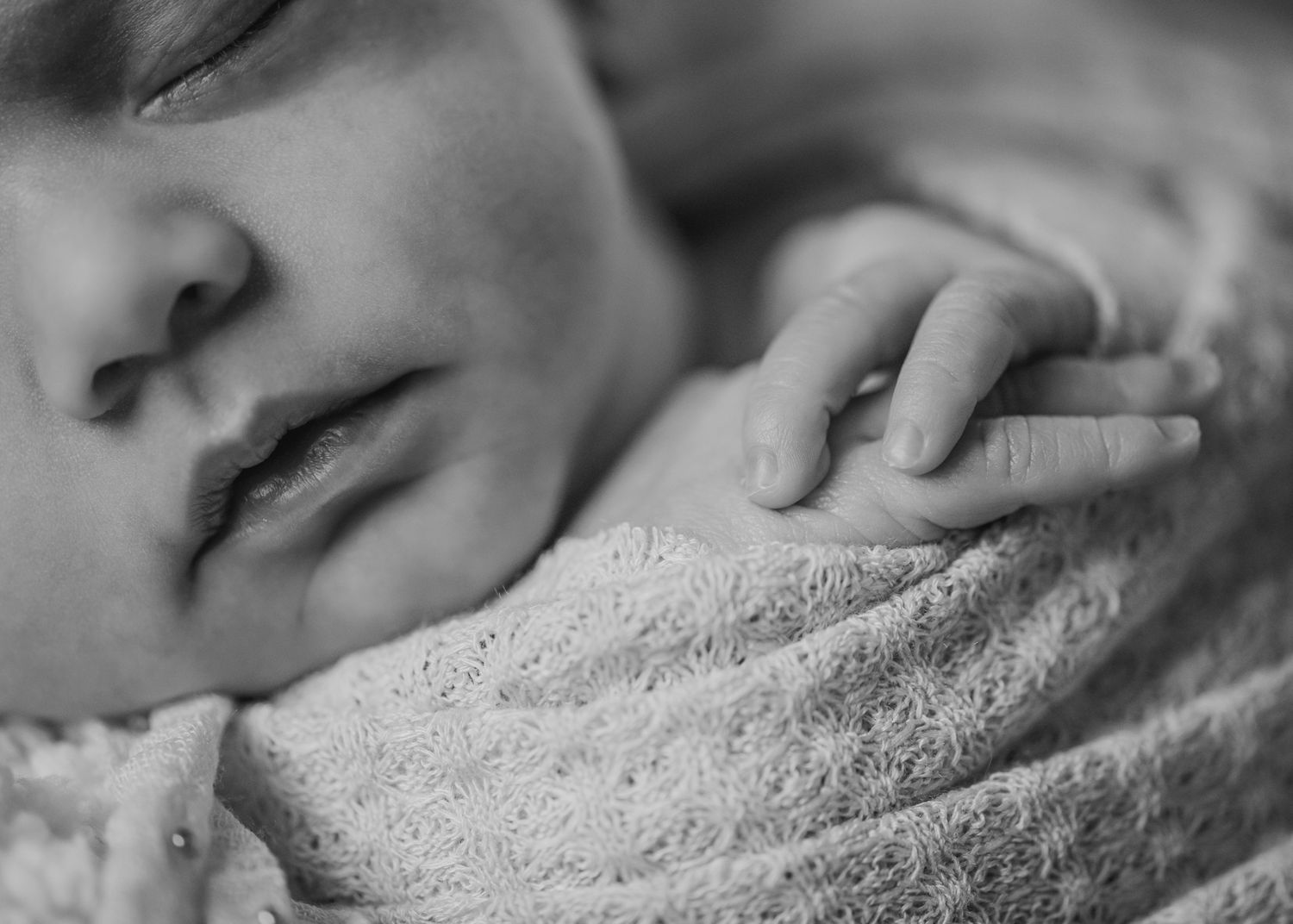 Close-up black and white photograph of a sleeping baby's face and hand resting on a soft blanket.