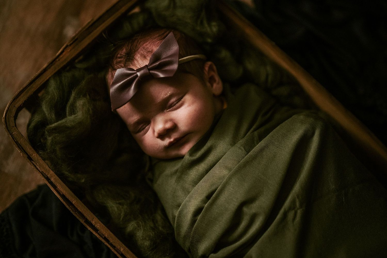 A sleeping baby wearing an olive green outfit and bow rests peacefully in a dark-toned bassinet.