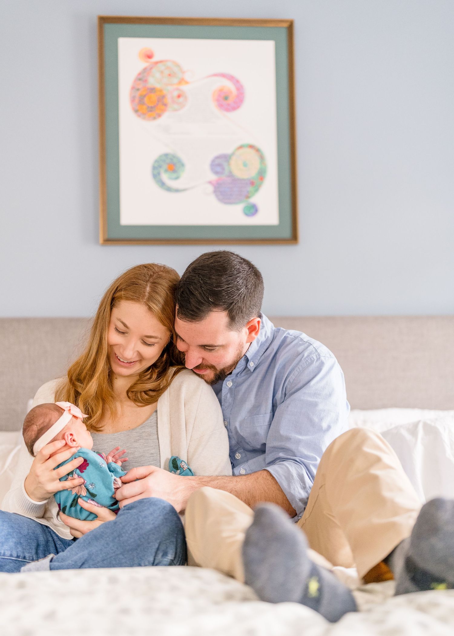 Parents cuddling with newborn on beige couch under framed wall art.