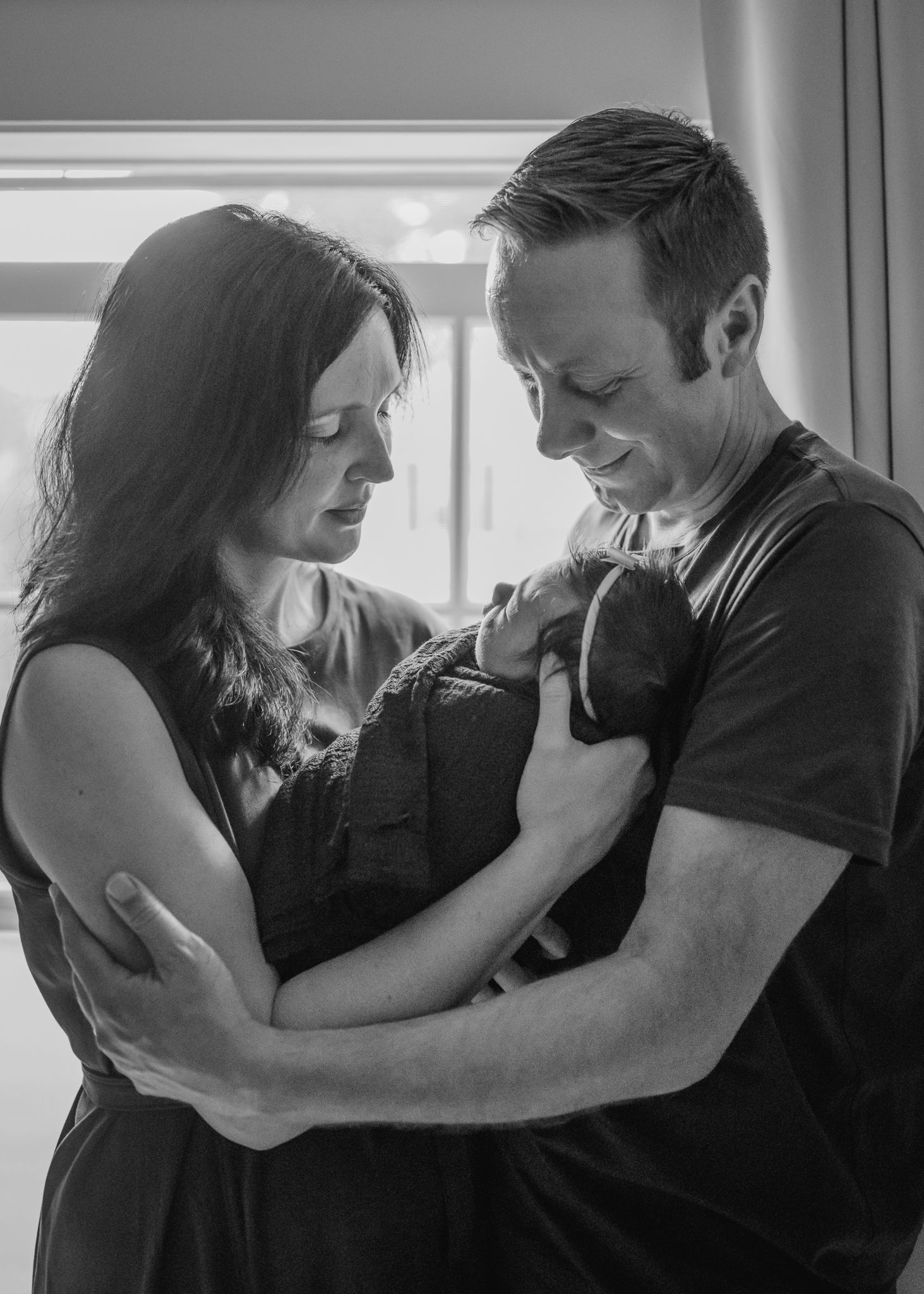 Black and white portrait of couple embracing while holding newborn baby by window.
