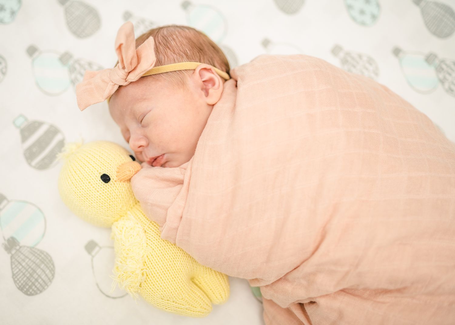 Sleeping newborn in peachy wrap with yellow plush duck on patterned background.