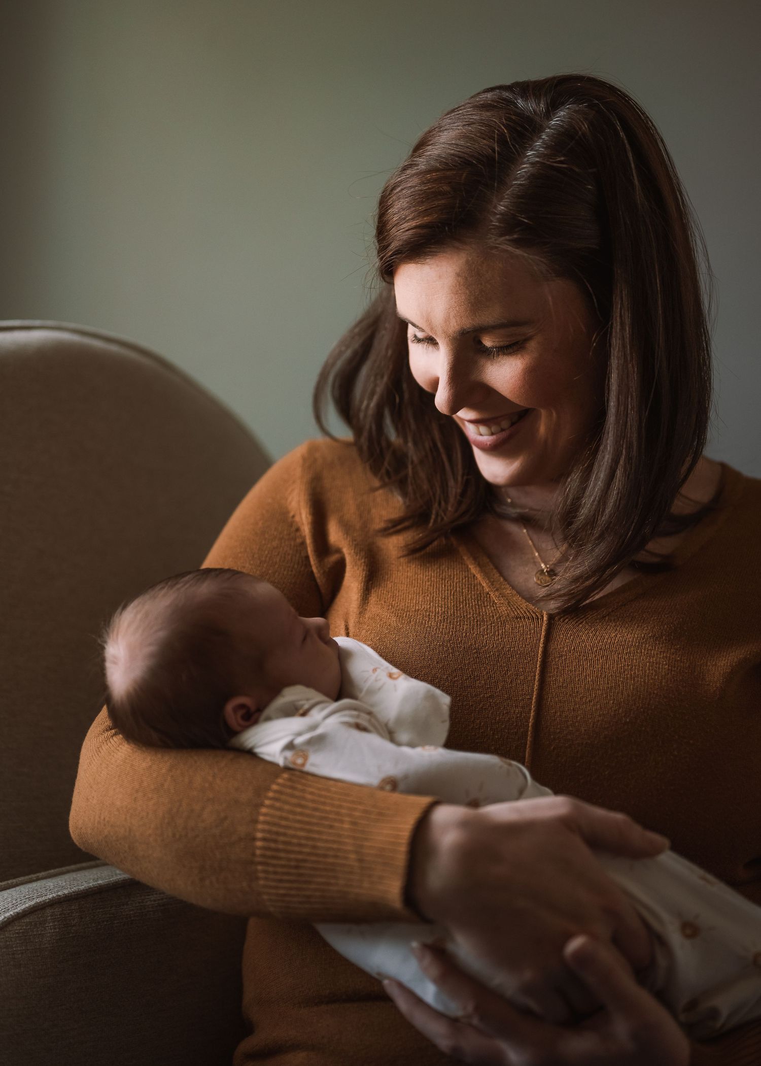 Mother in brown sweater holds sleeping infant while sitting in plush gray chair.
