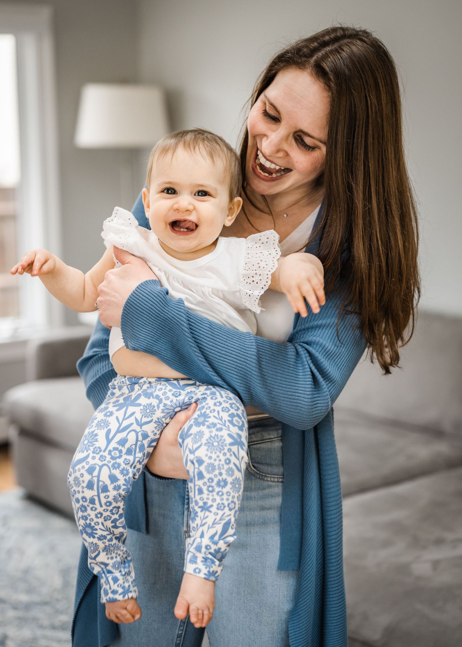 A joyful moment in a living room as a baby in white lace and blue floral pants is held by an adult in a blue cardigan.
