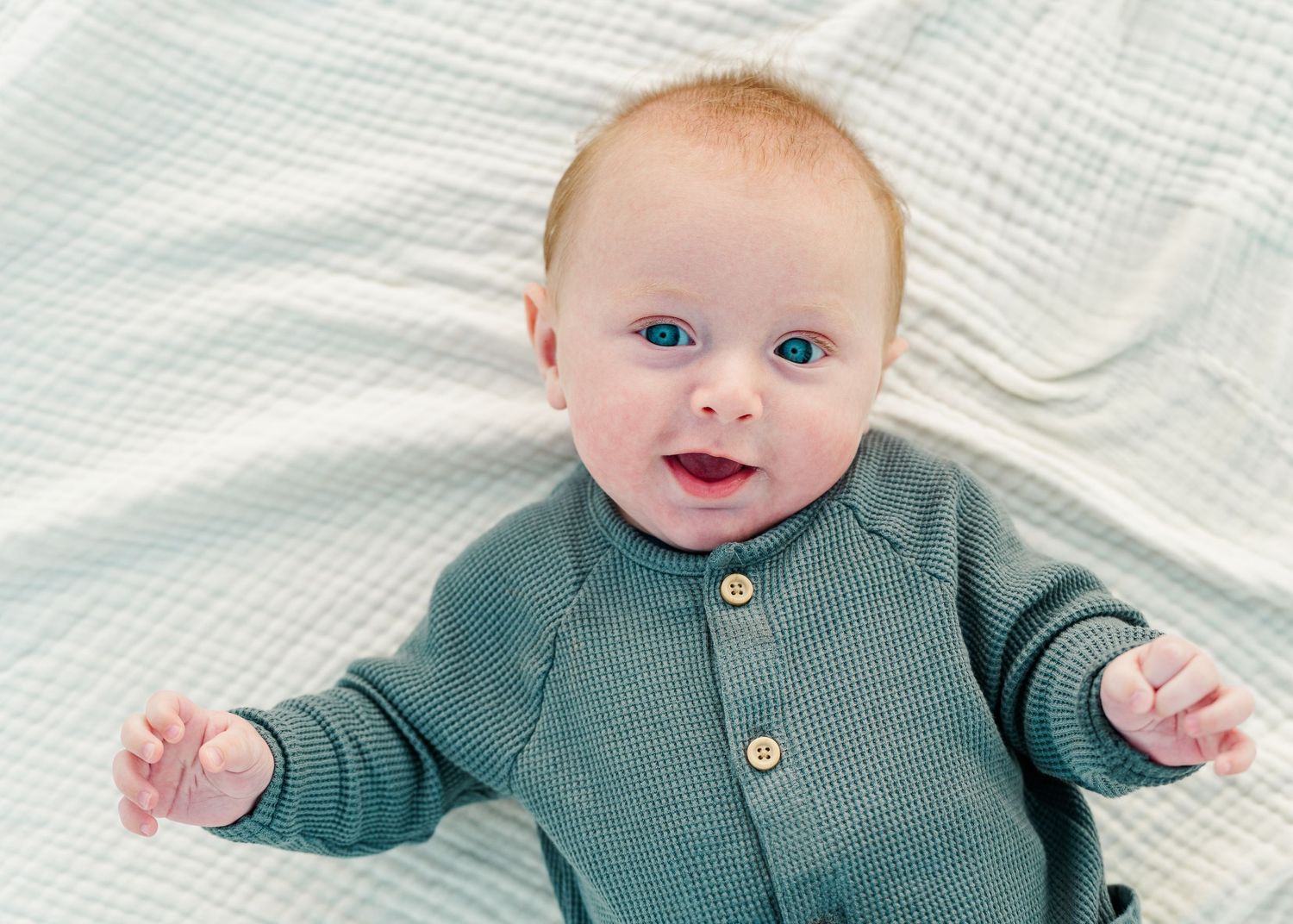 Smiling baby wearing teal cardigan sweater lying on white textured blanket.