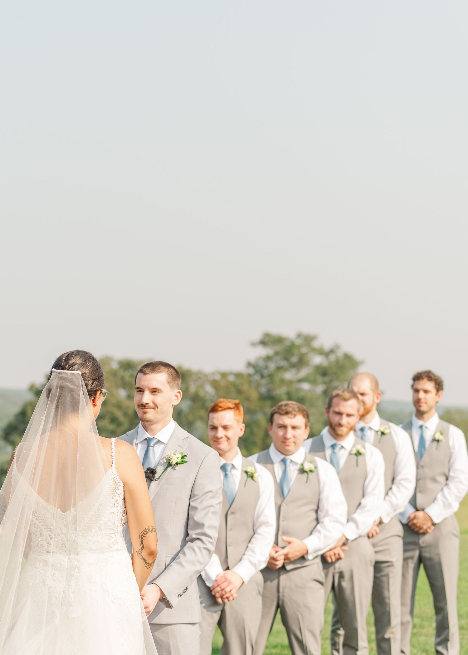 Wedding party in light gray suits stands in a row on grassy field with bride in white gown and veil at sunset.