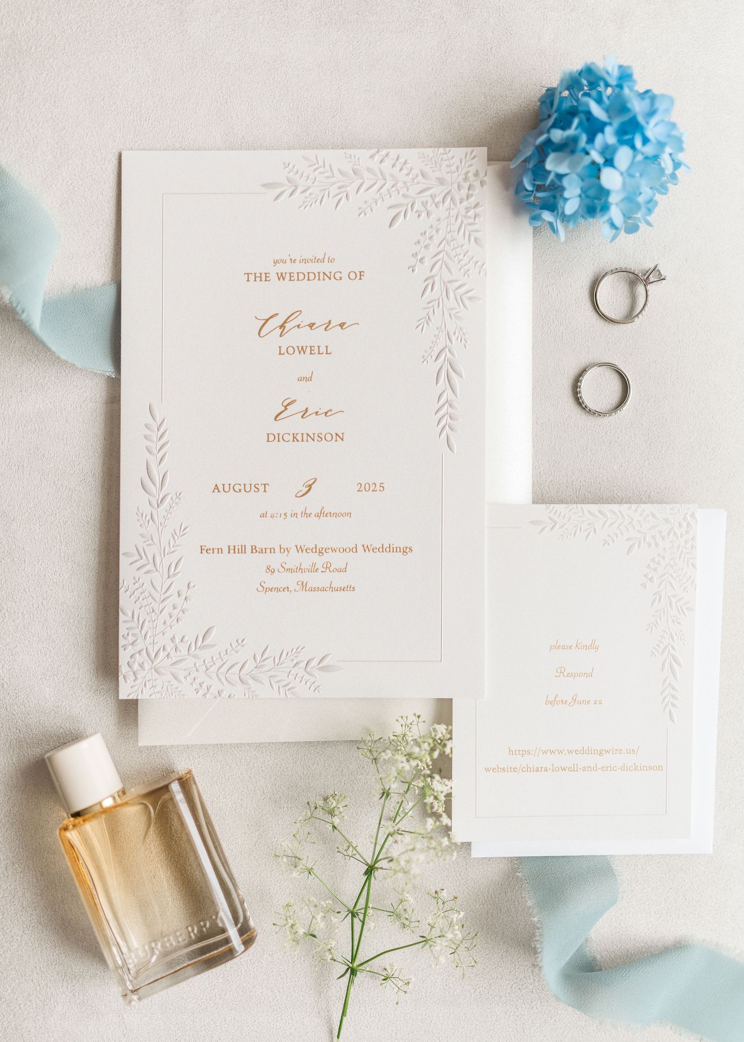 Elegant wedding stationery with blue flowers, perfume bottle, and delicate white floral embossed details arranged on light surface.