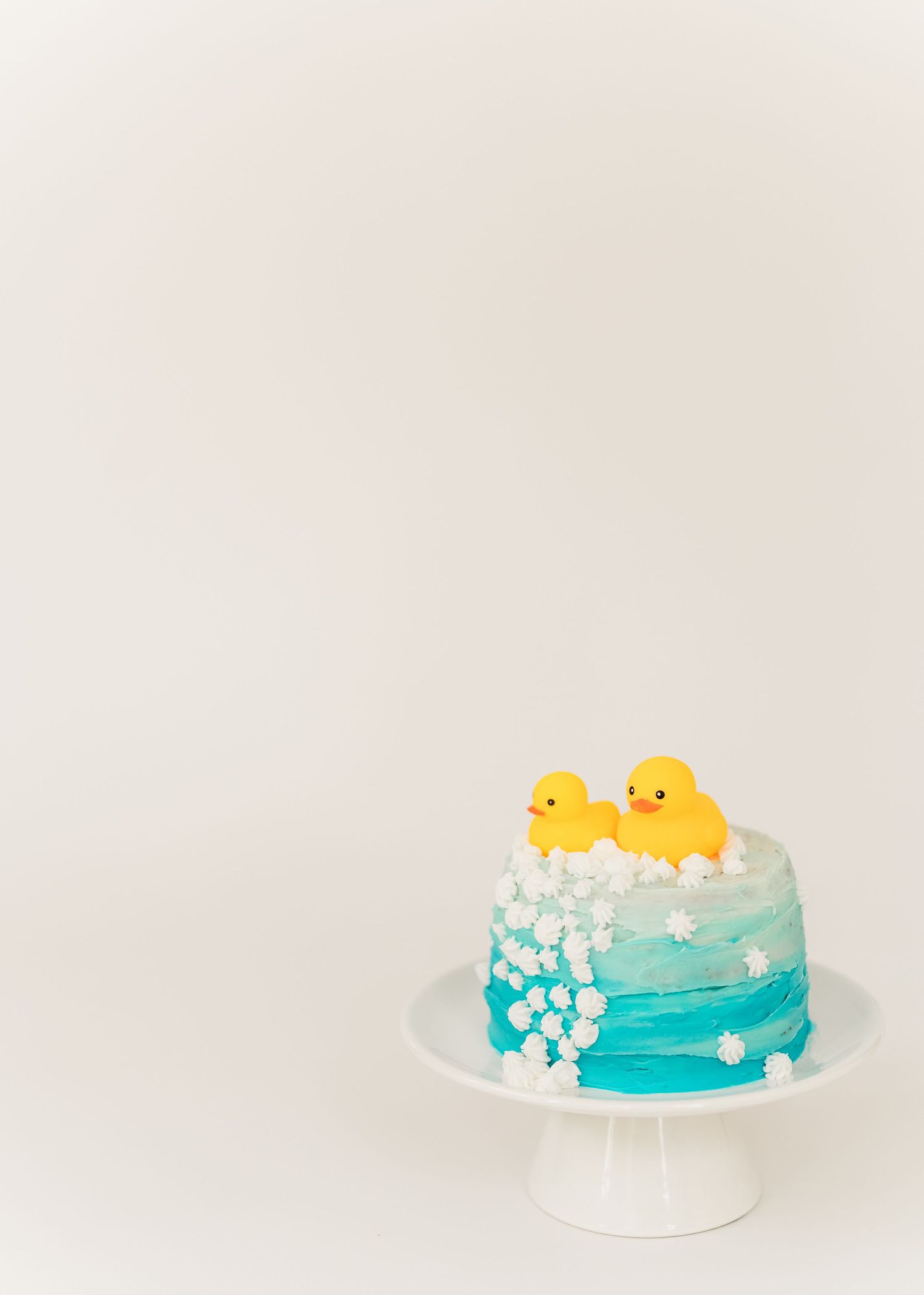 Cake smash. A turquoise ombre cake decorated with white frosting and topped with two yellow rubber ducks sits on a white cake stand.