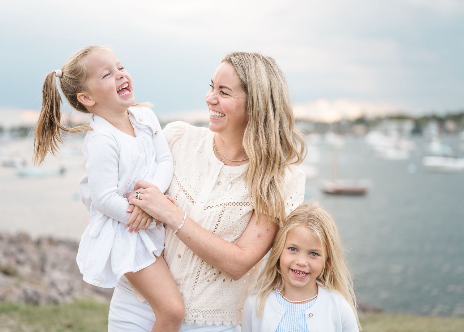 A joyful family shares laughter and embraces while wearing white outfits during an outdoor photoshoot near the water.