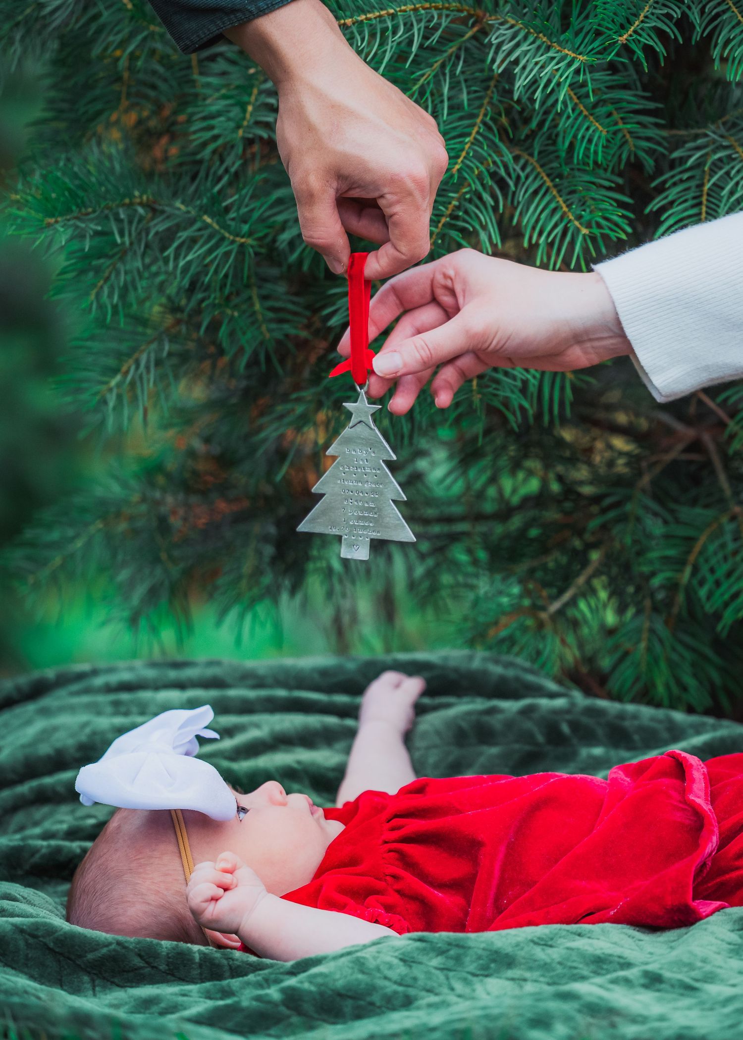 Hands hold Christmas tree ornament above baby lying on green velvet blanket.