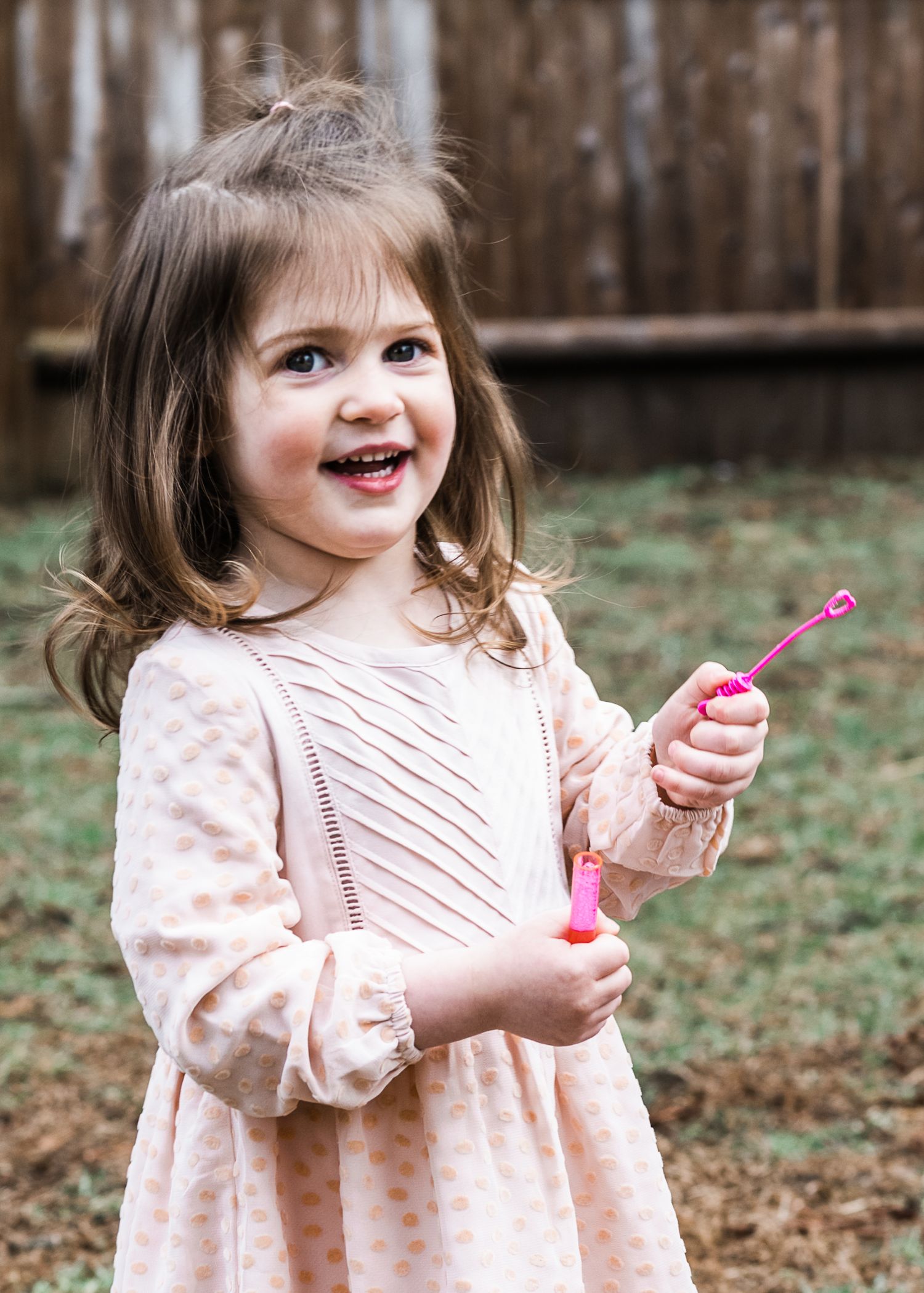 A cheerful child in a pink patterned dress holding a bubble wand outdoors.