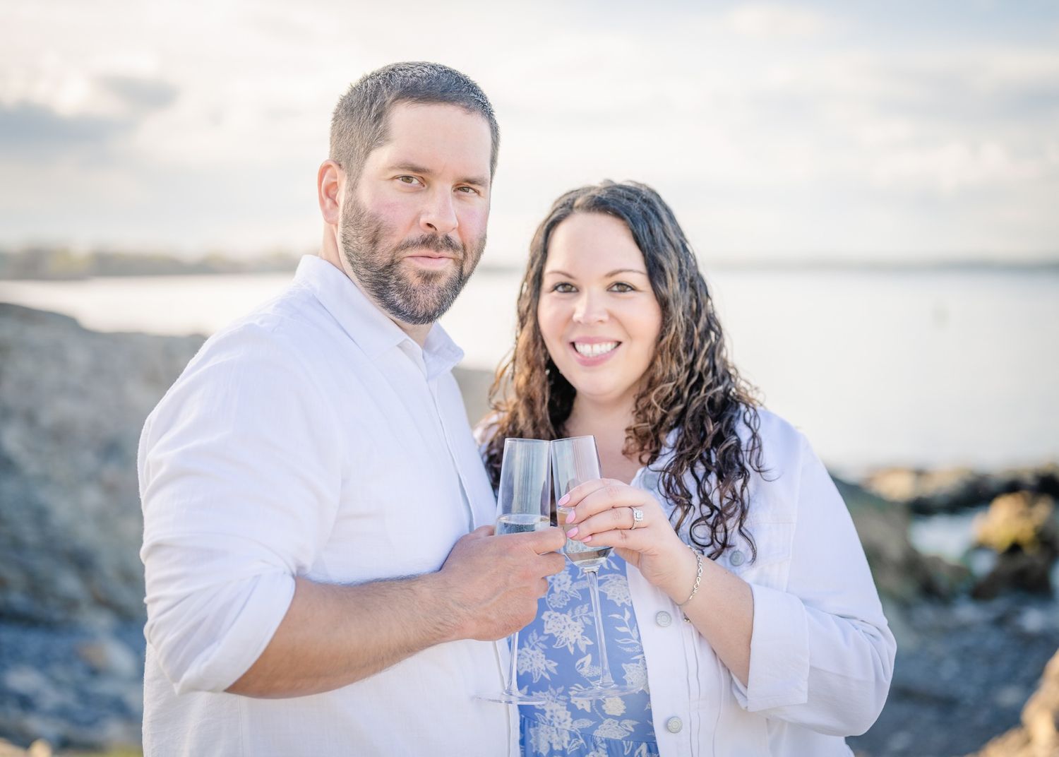 A couple shares a toast with wine glasses against a coastal backdrop at sunset.