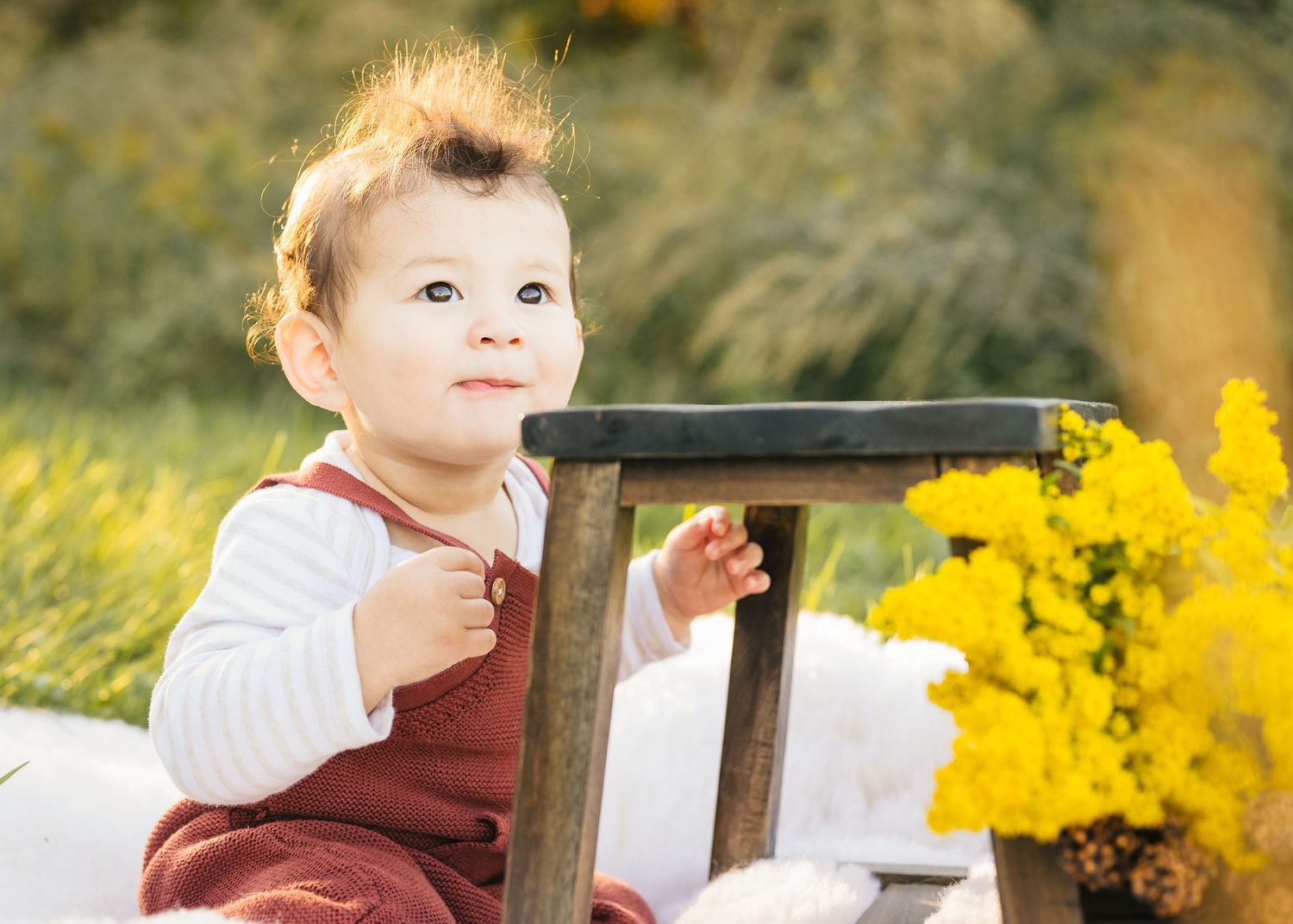 A baby in burgundy overalls sits near yellow flowers and a wooden chair in a garden setting.
