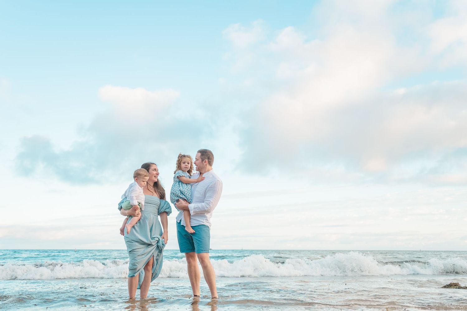 A family wearing light blue outfits stands together at the beach during sunset with waves rolling in behind them.