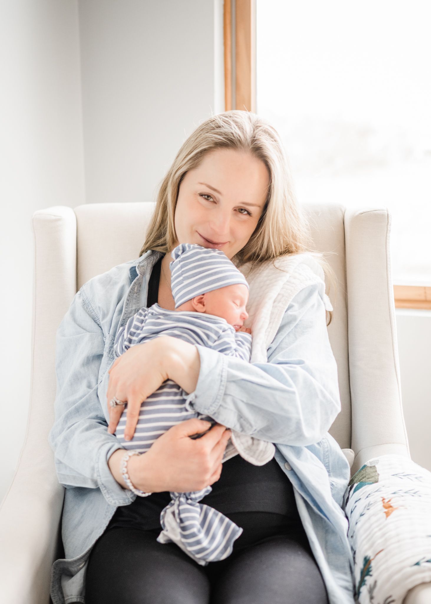 Tender moment in white armchair with sleeping infant in grey stripes and denim.