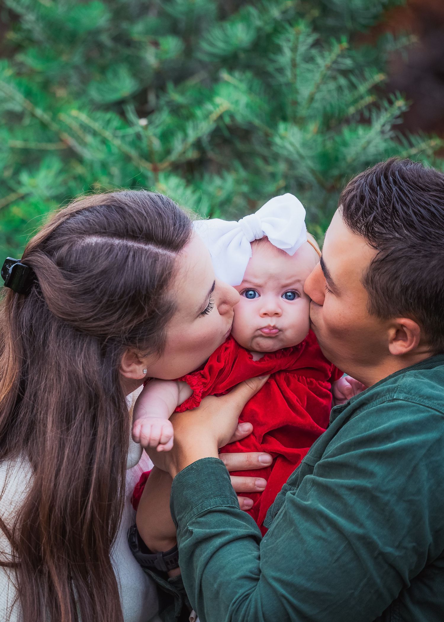 Sweet holiday moment with baby in red dress surrounded by evergreen branches.