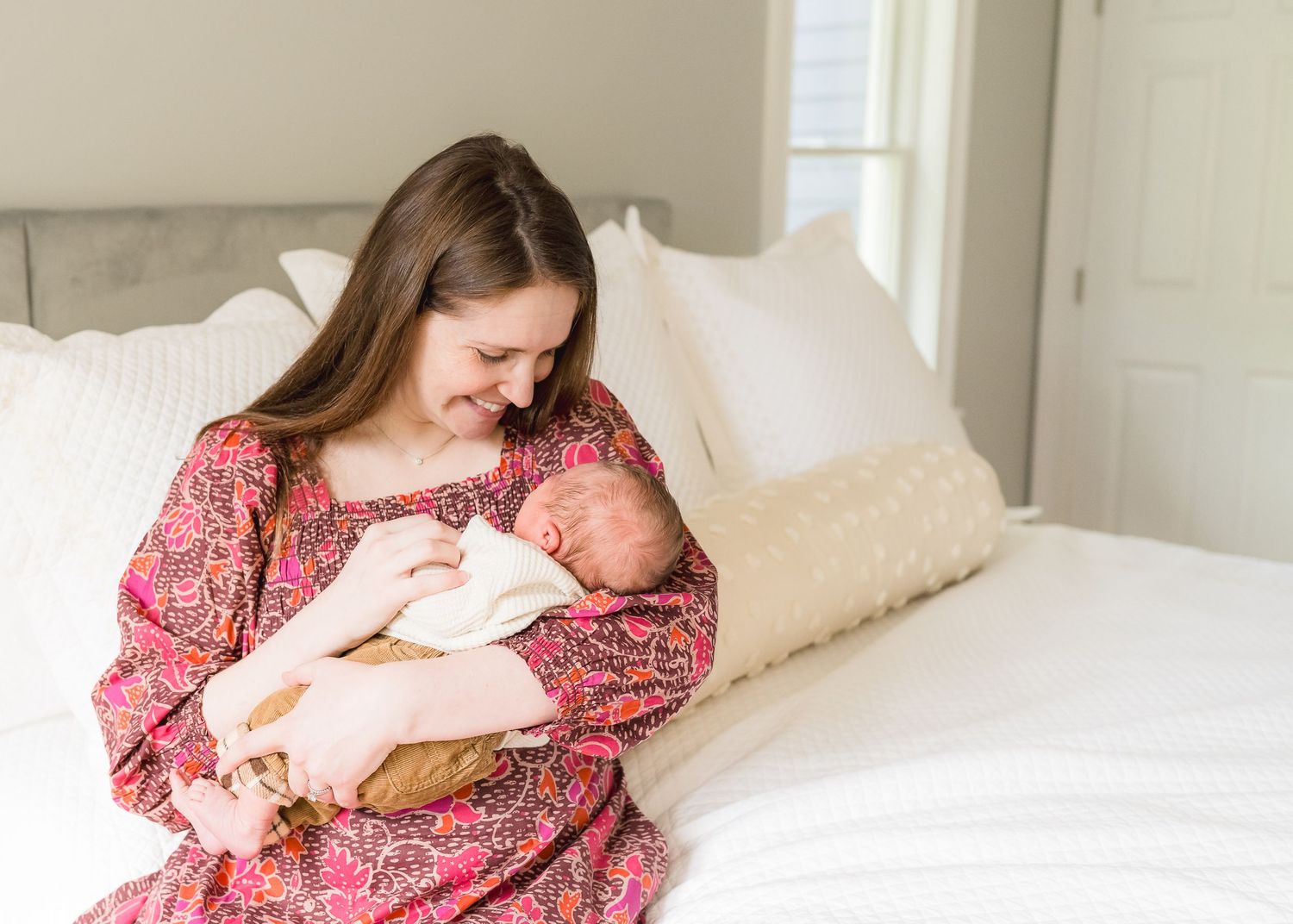 Mother in floral dress cradling newborn baby on white bed.