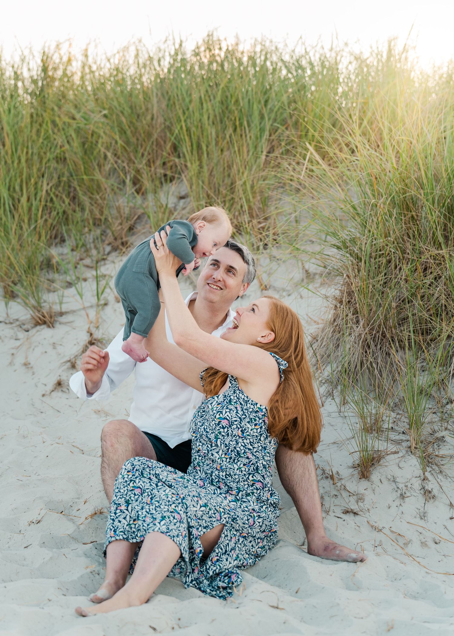 A loving moment captured between a family playing together on a sandy beach surrounded by dune grass at sunset.