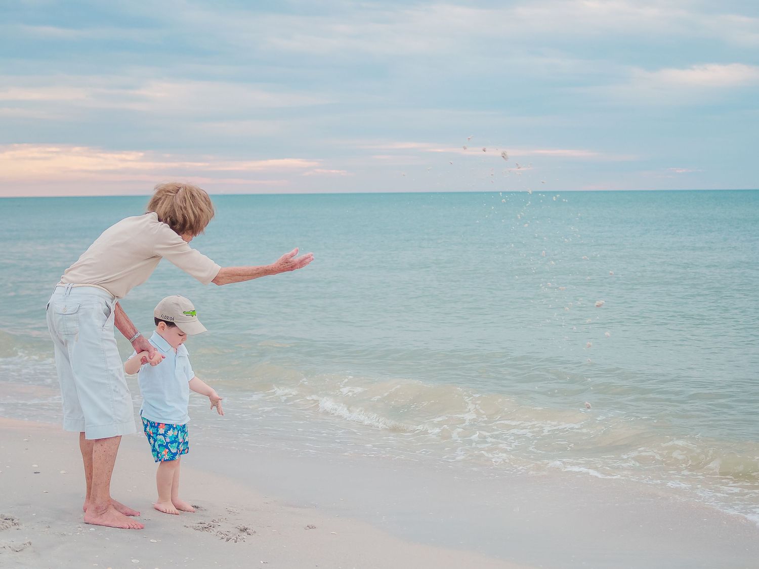 Parent and small child stand at the edge of calm ocean waters pointing at something in the distance on a peaceful beach.