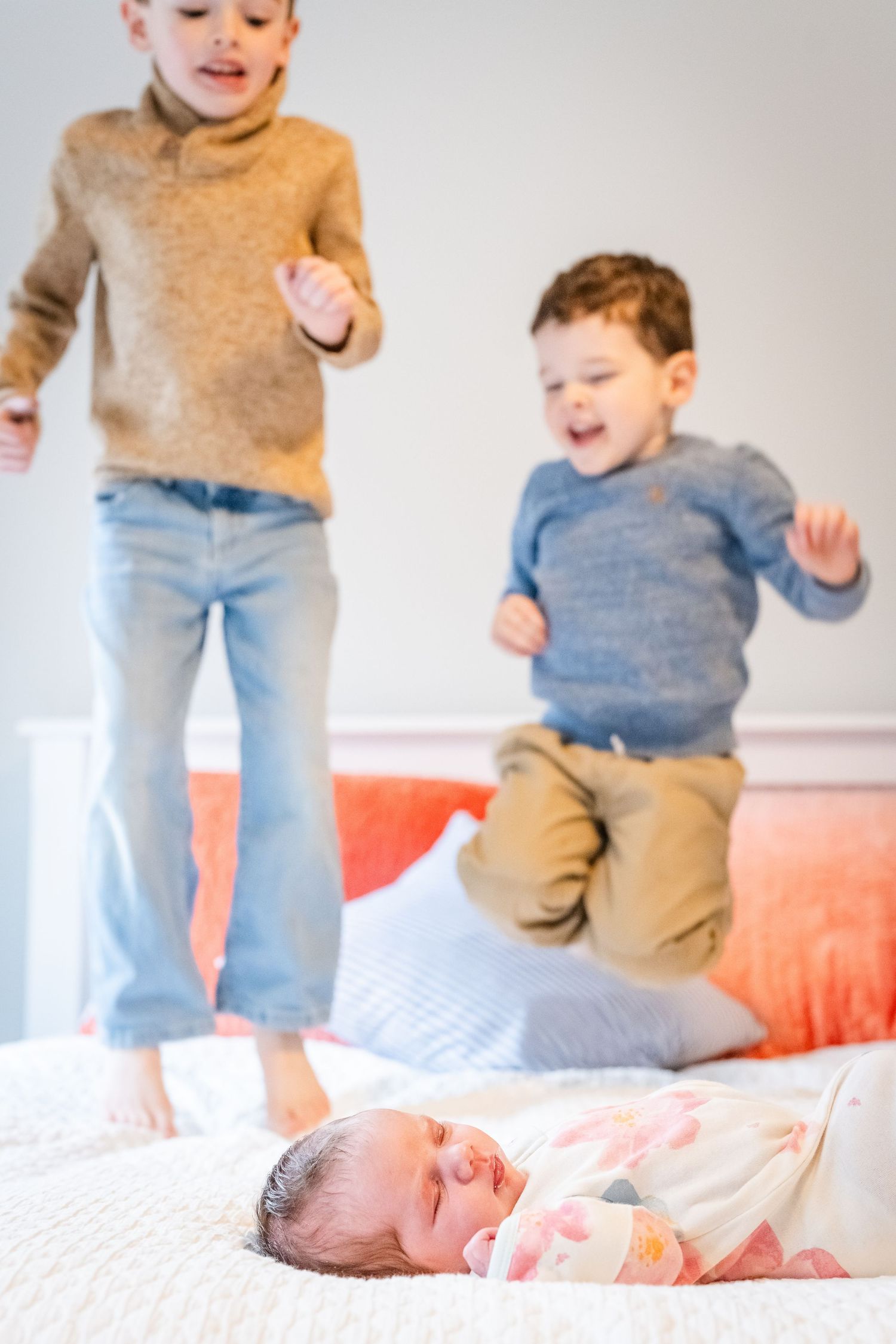Two children jumping on a bed while a baby in pink sleeps peacefully below them.