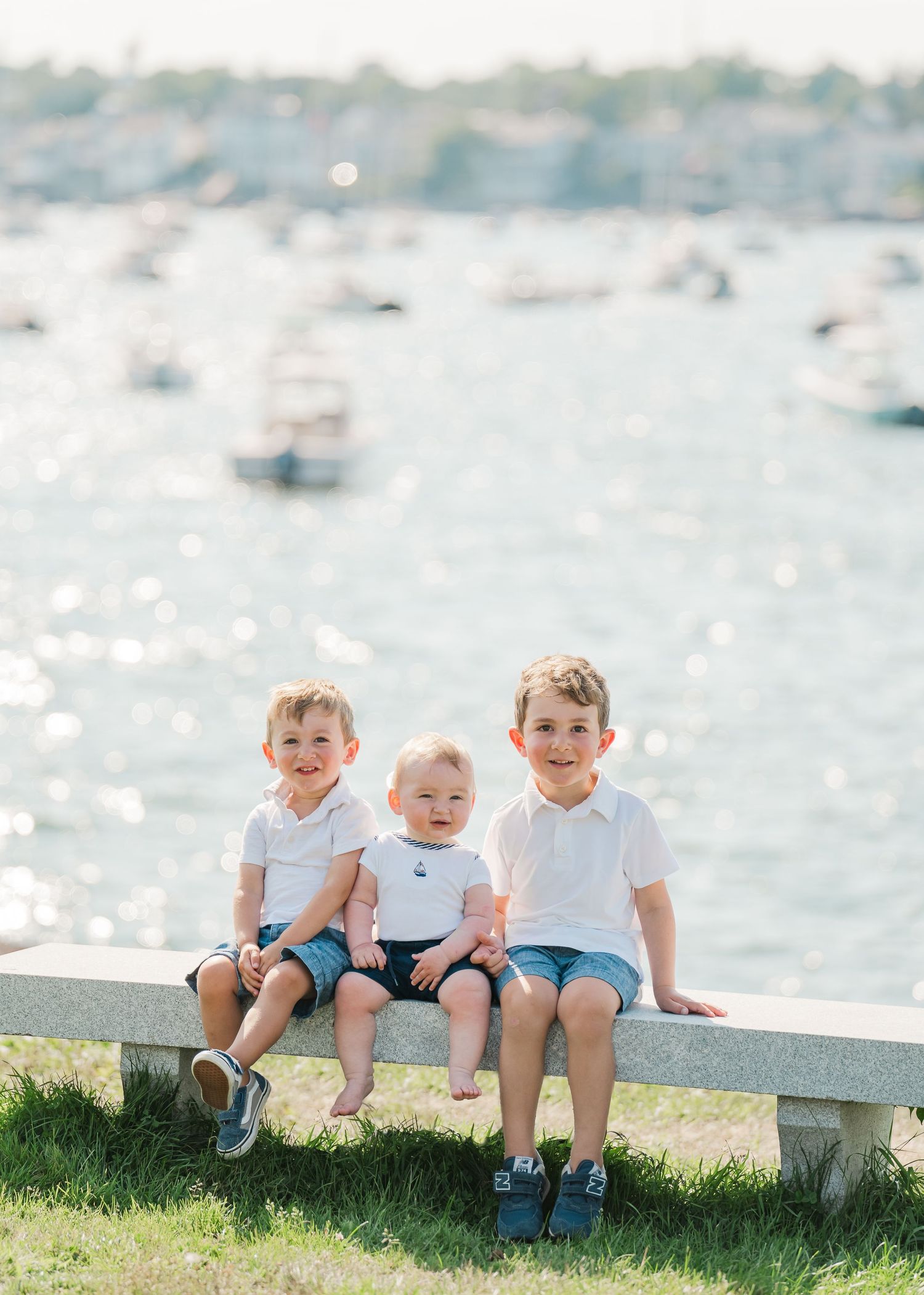 Three young children sitting together on a stone bench by the sparkling water on a sunny summer day.