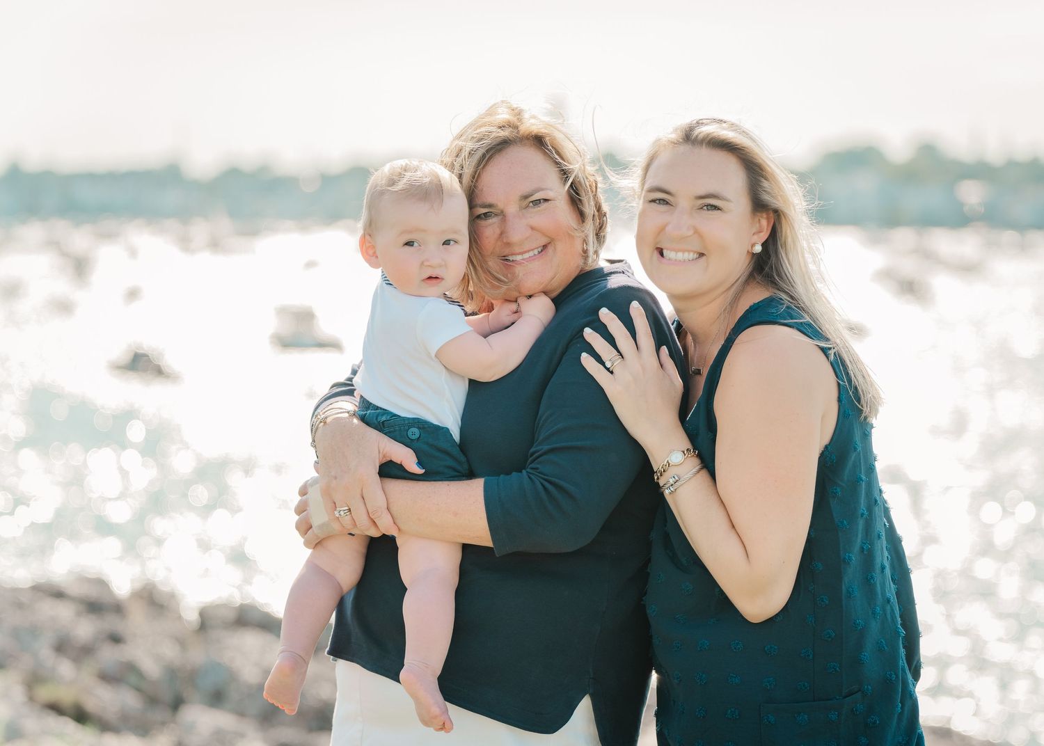 A family poses together on a rocky beach against sparkling water on a sunny afternoon.