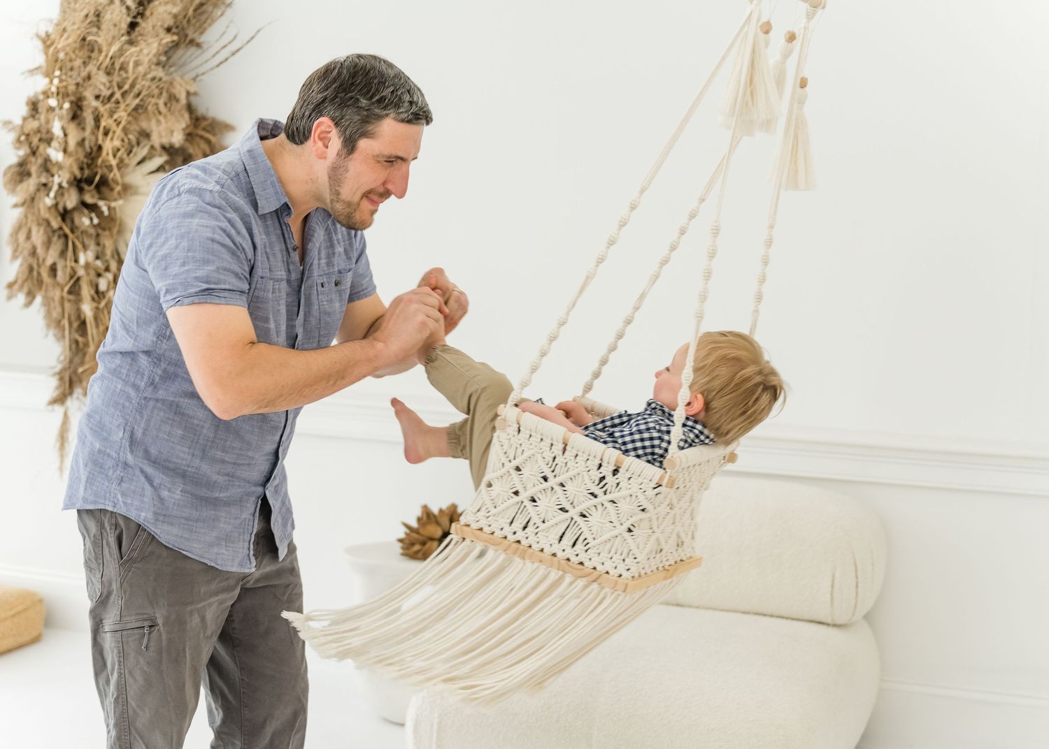 Parent gently pushing a toddler in a macrame baby swing inside a bright white modern home.