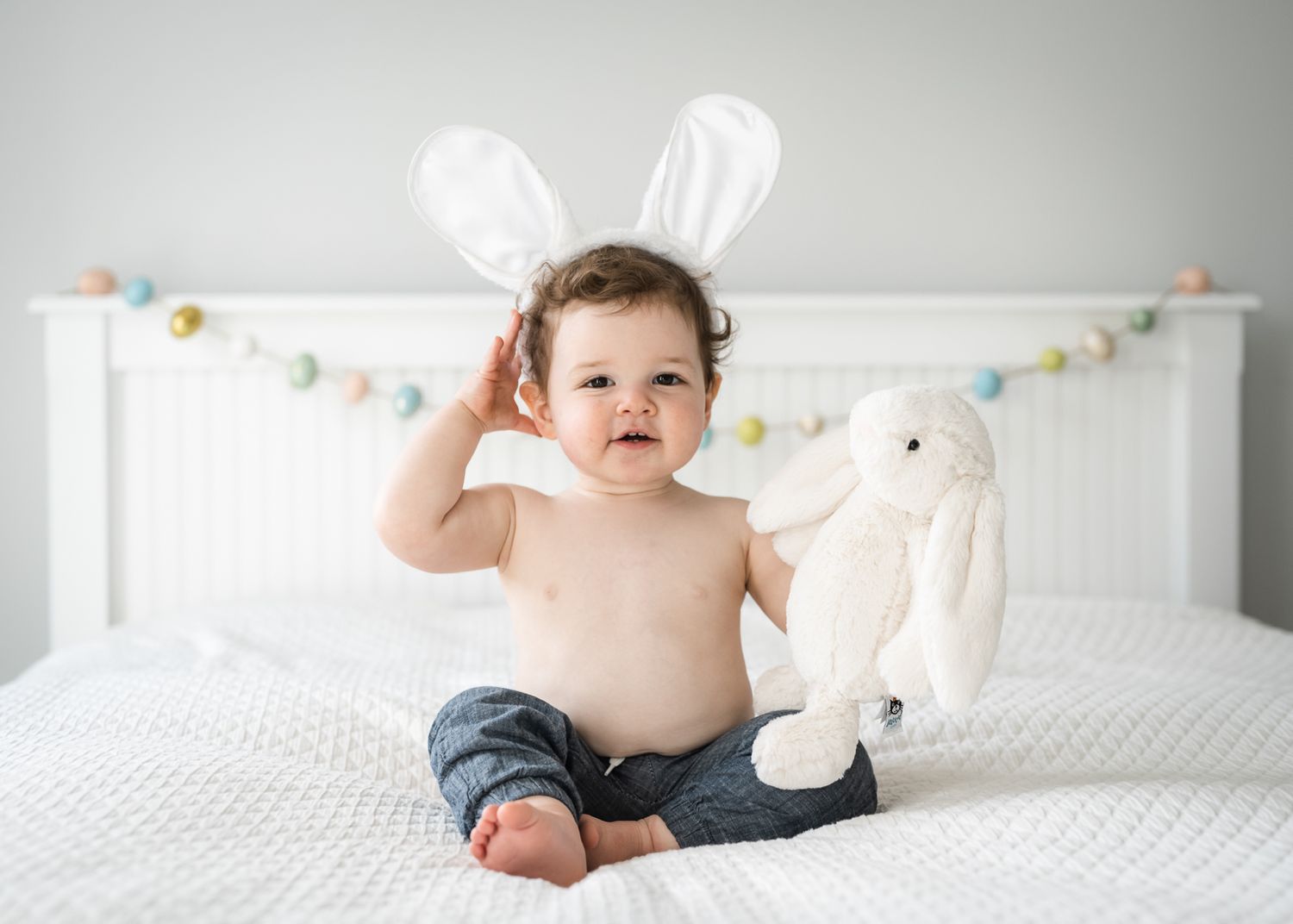 Baby wearing bunny ears sits on white bed with stuffed rabbit toy.