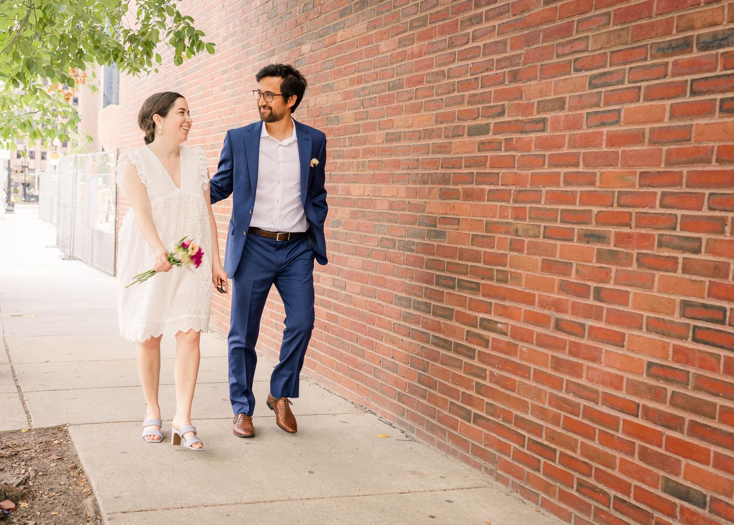 Couple walking together along brick wall sidewalk in formal attire.
