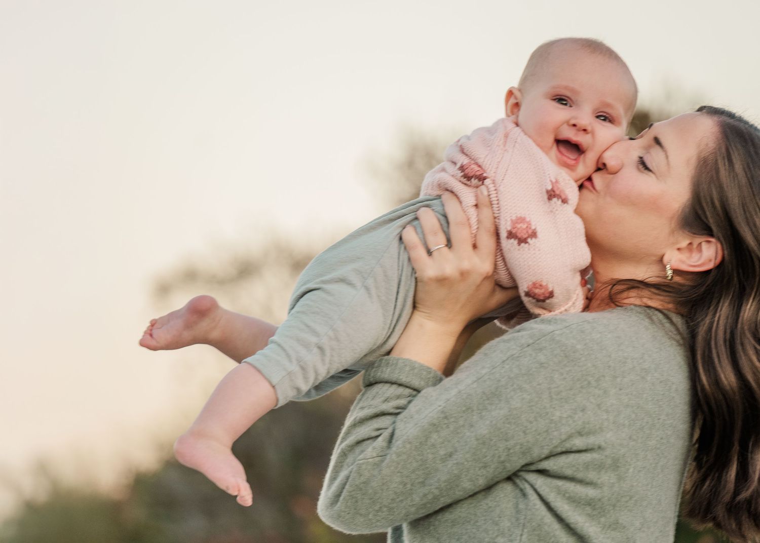A parent in a gray sweater lifts and plays with a joyful baby against a bright outdoor background.