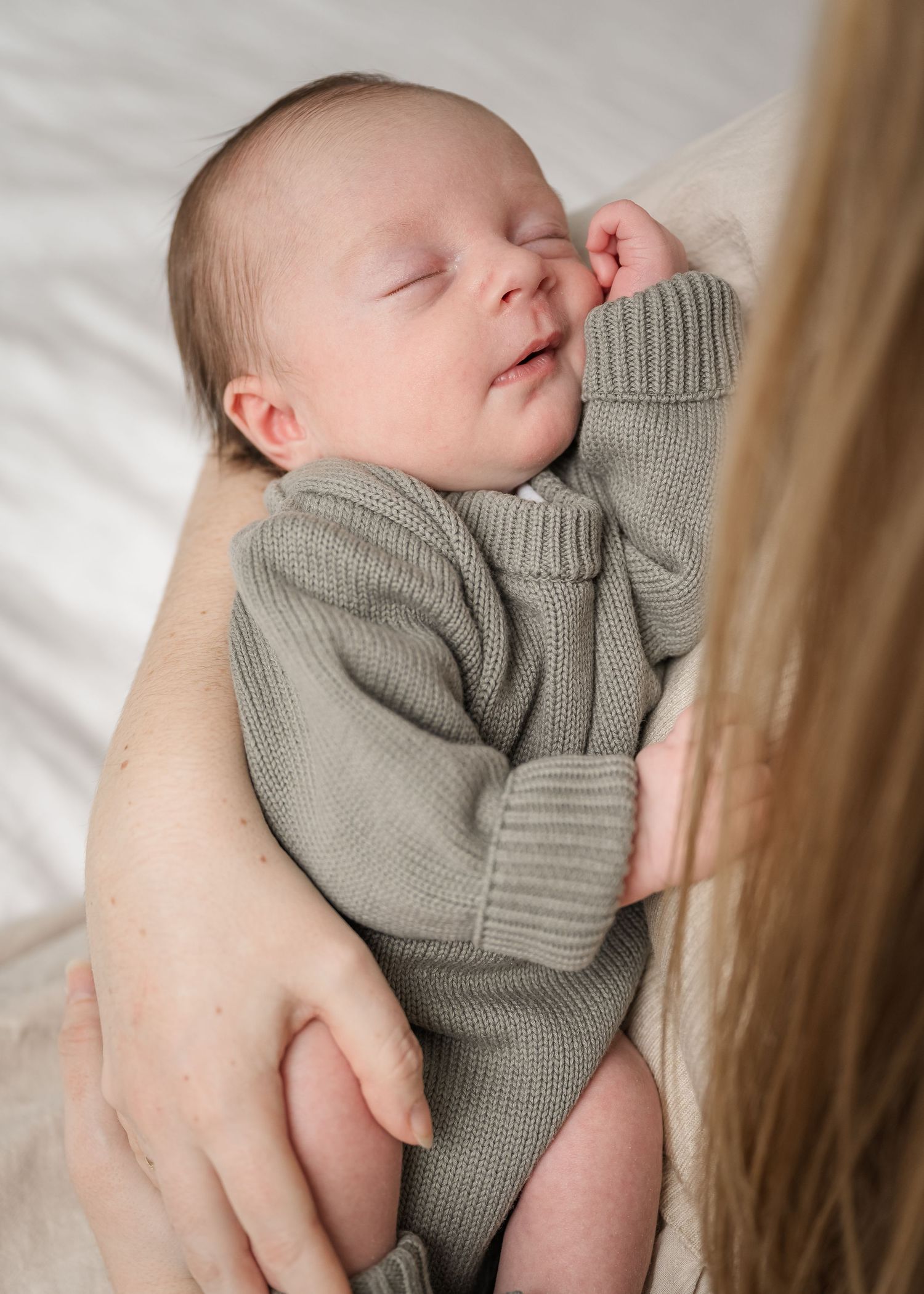 Peaceful sleeping newborn baby wearing a cozy gray knit sweater rests in gentle arms.