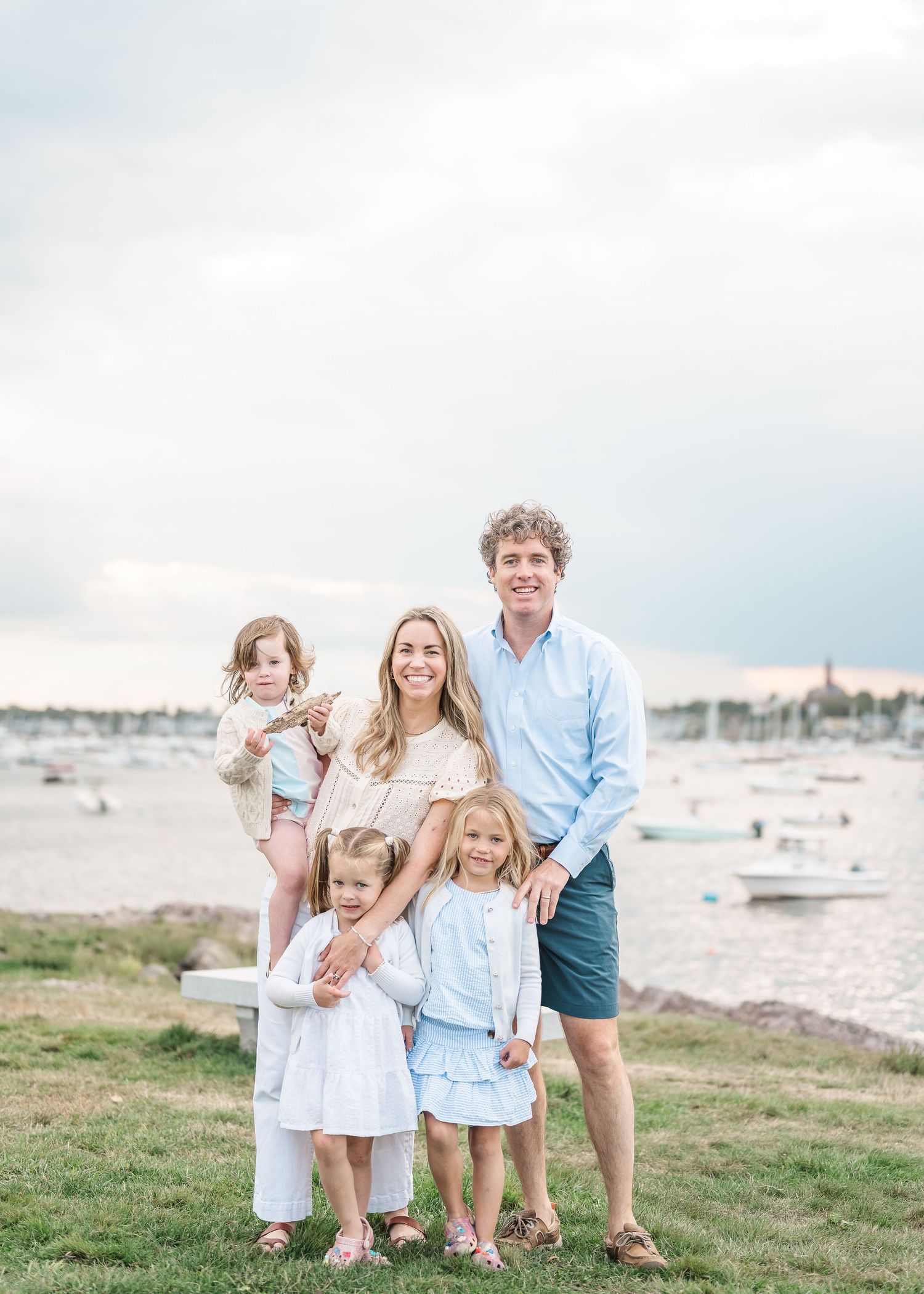 A family stands together on grassy coastline with boats and harbor in background at sunset.