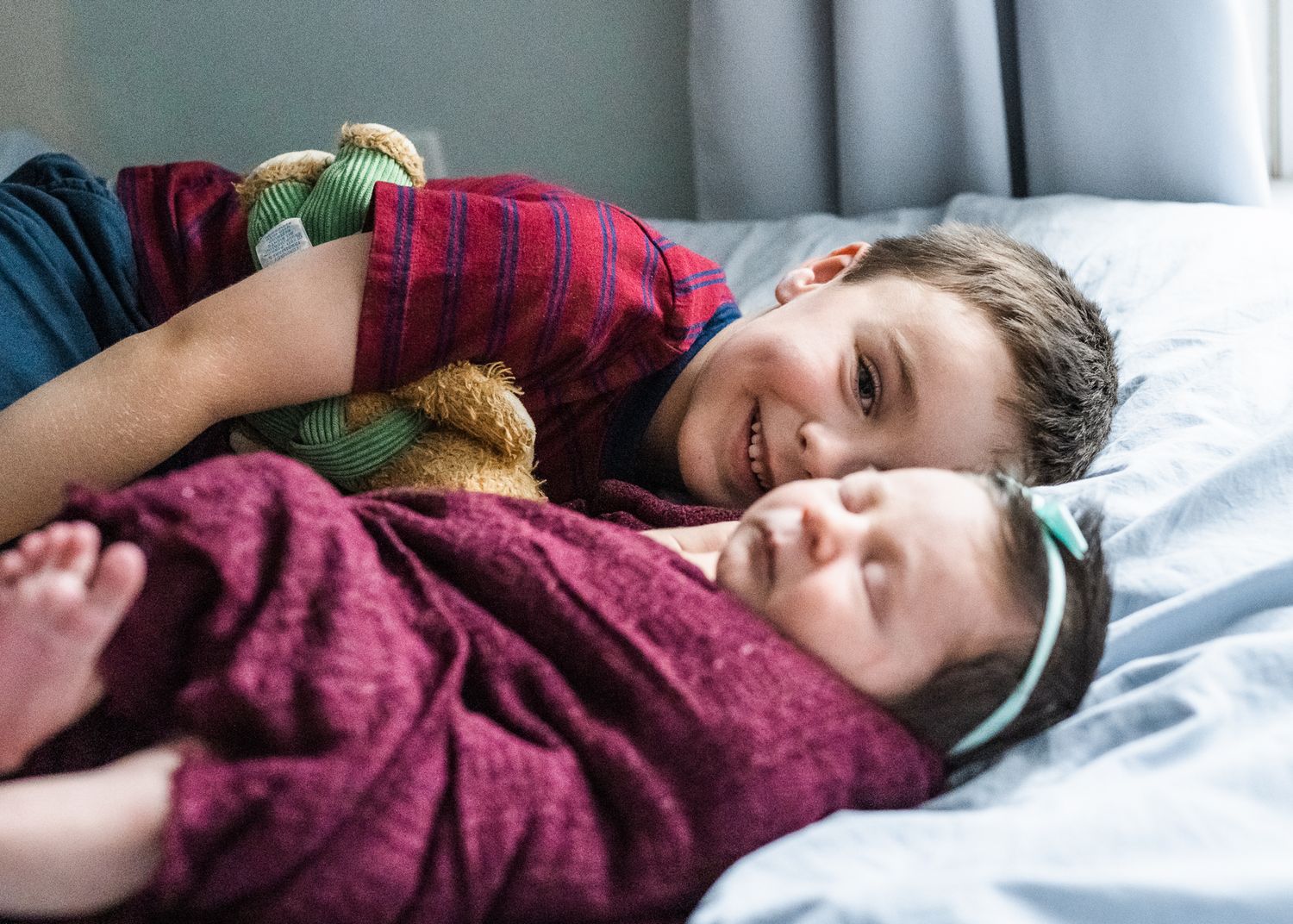 Two siblings sharing tender moment while resting together on bed with stuffed toy.
