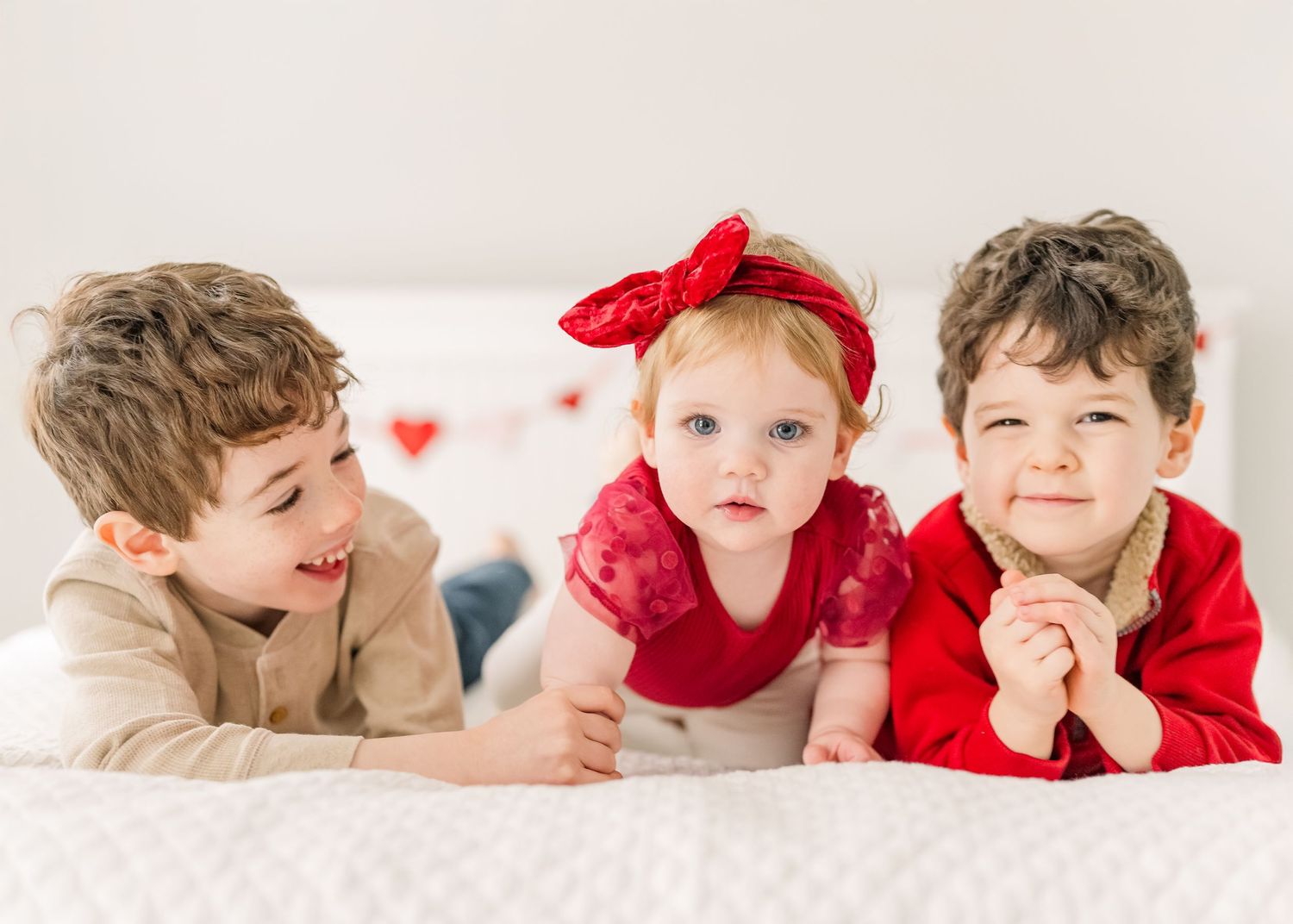 Three children in red and white holiday outfits lying together on soft white bedding for a portrait session.