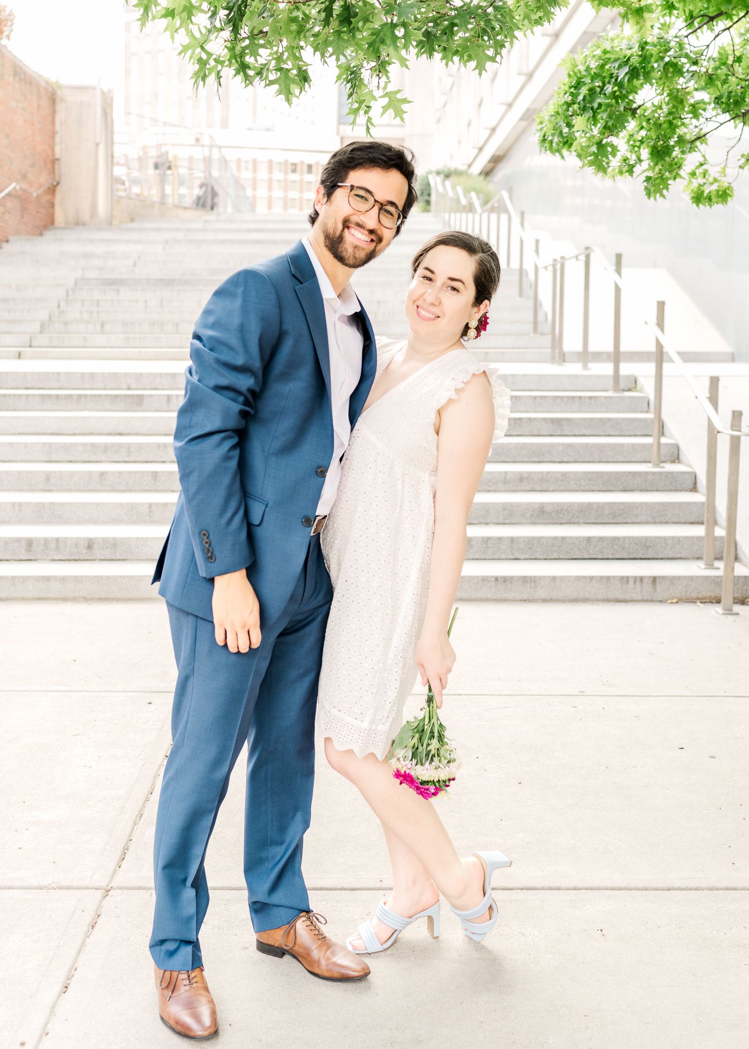 Couple poses on bright outdoor stairway with greenery overhead.