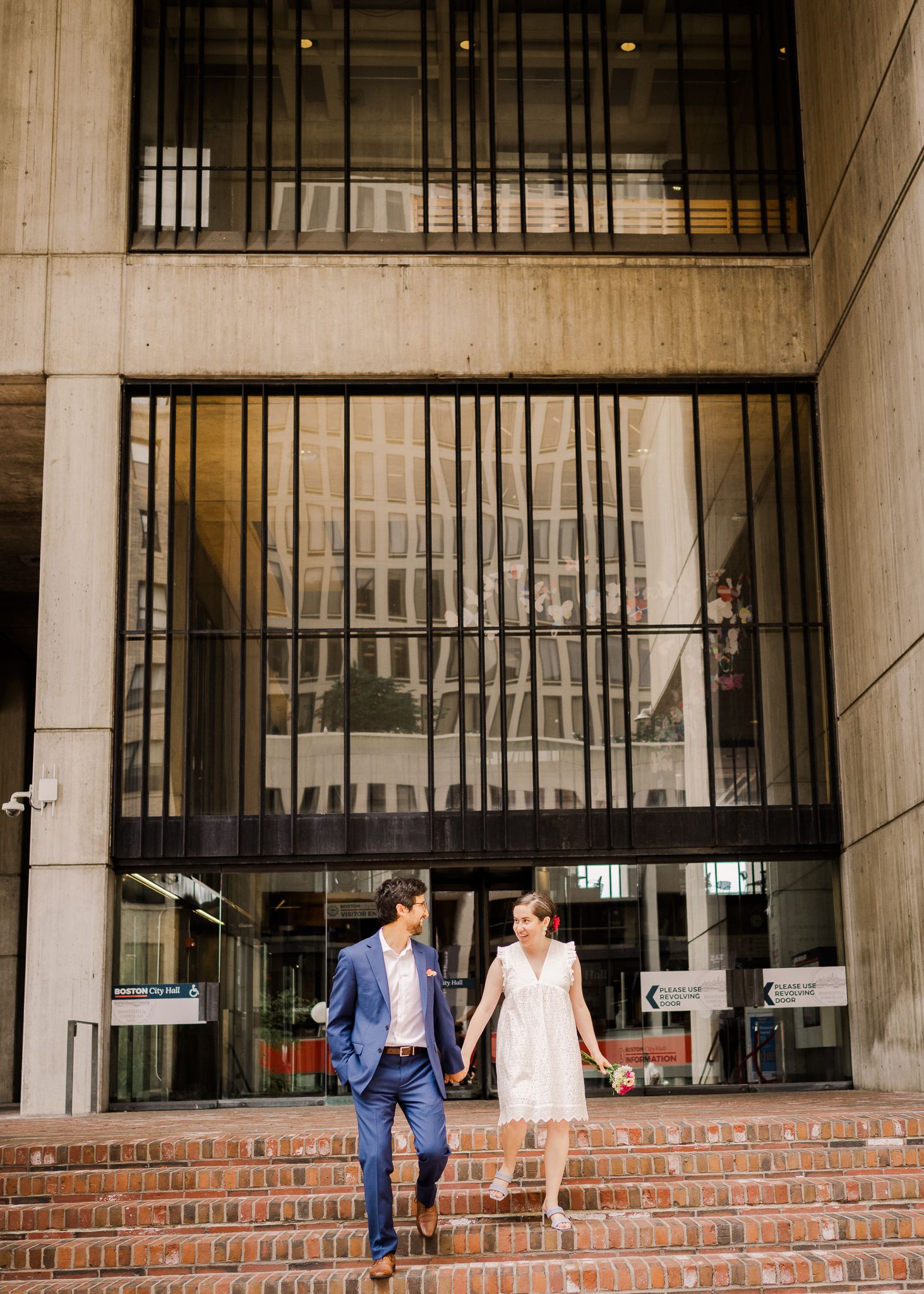 A couple walks down brick steps in front of a modern glass building facade.