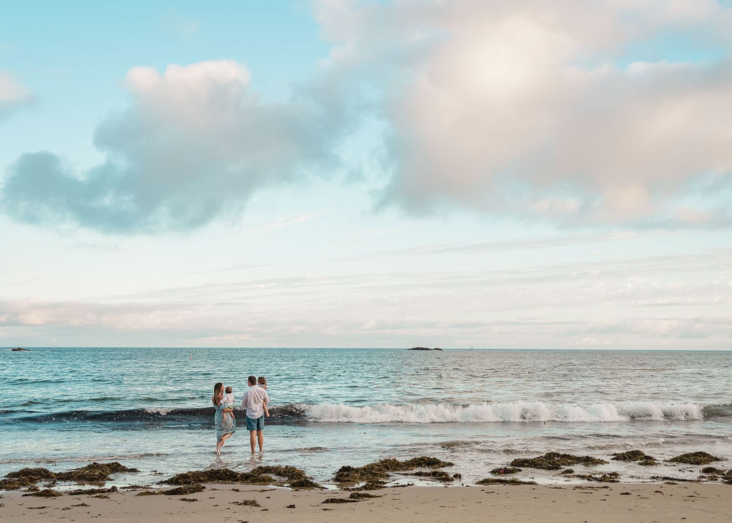 Two people stand together on a rocky beach at sunset with turquoise ocean waters and soft clouds in the sky.