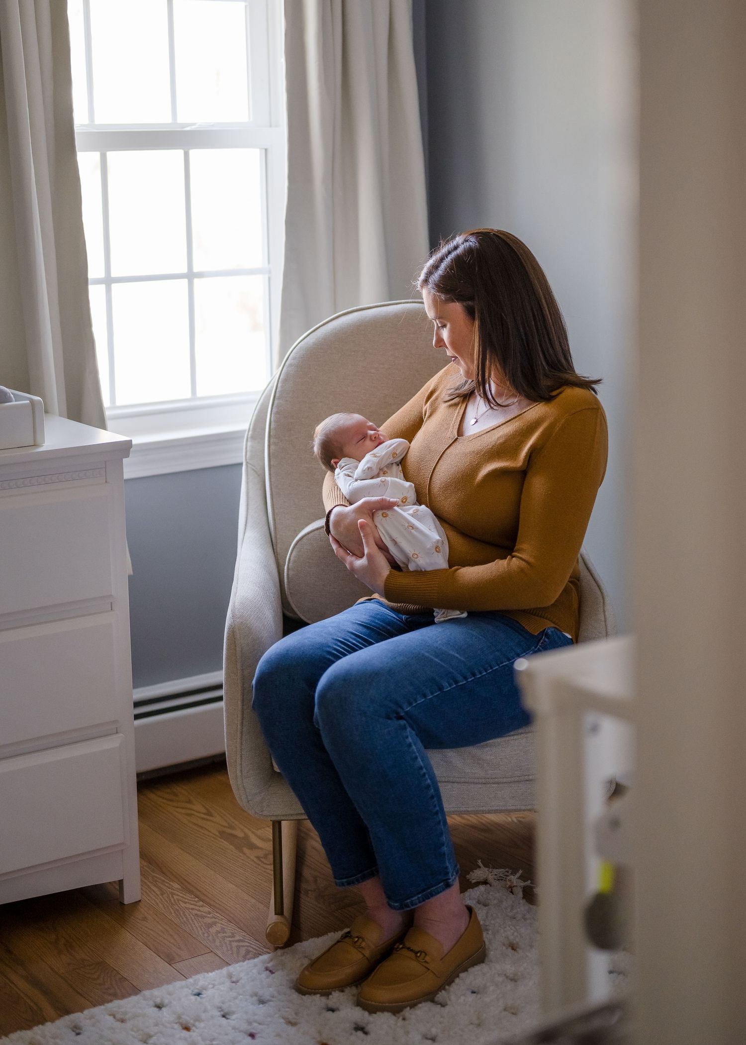 Mother in mustard sweater sits in nursery chair holding sleeping baby near window.