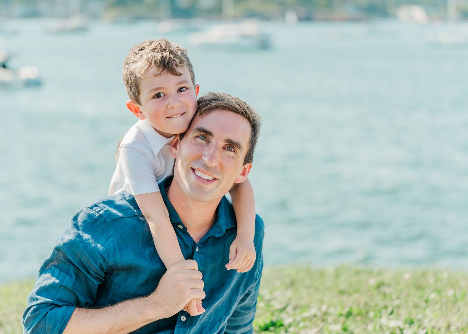 Parent and child enjoying a sunny day by the water with boats visible in the background.