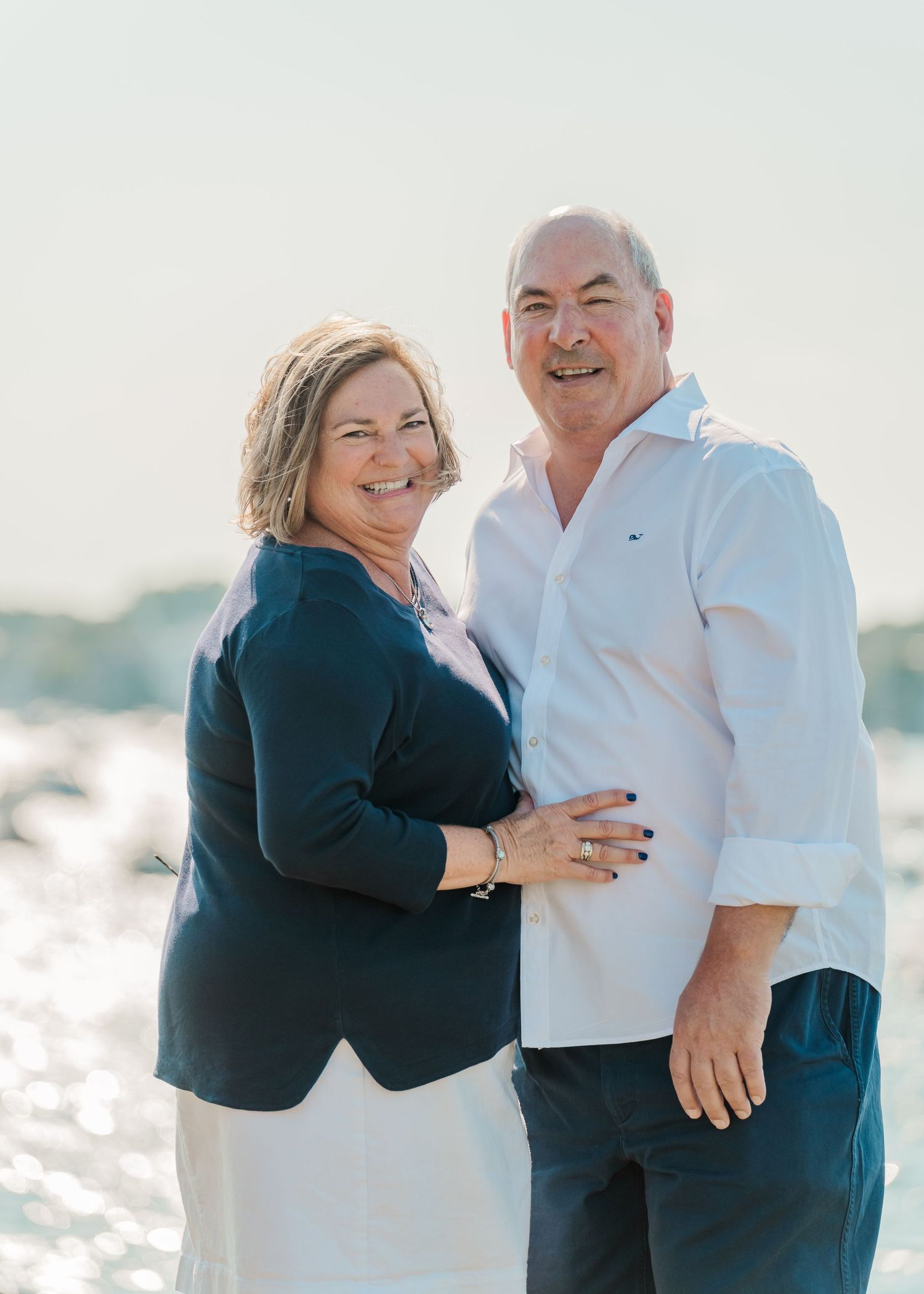 Couple in casual attire smiling together for a portrait by the water on a sunny day.