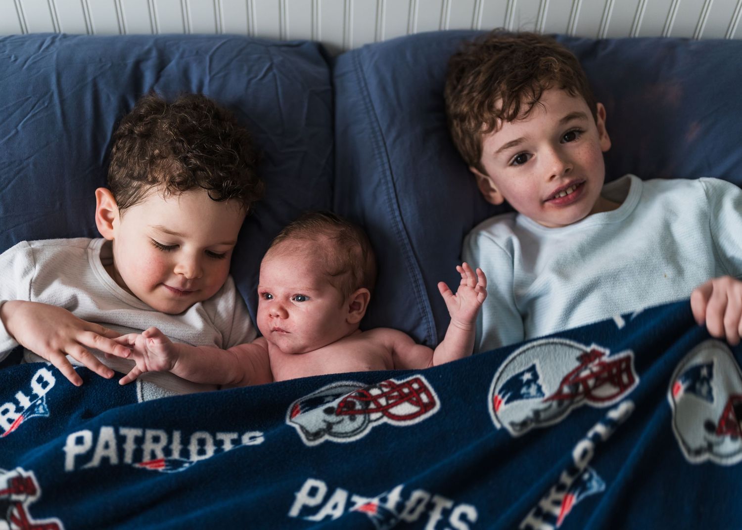 Three children cuddling together on a blue couch with Patriots NFL team blanket.