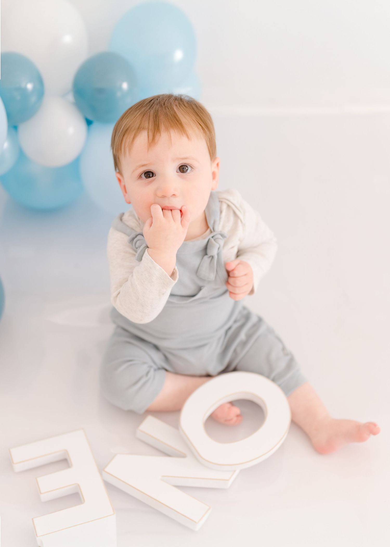 A small child in gray overalls sits on a white floor with decorative letters and blue balloons in the background.