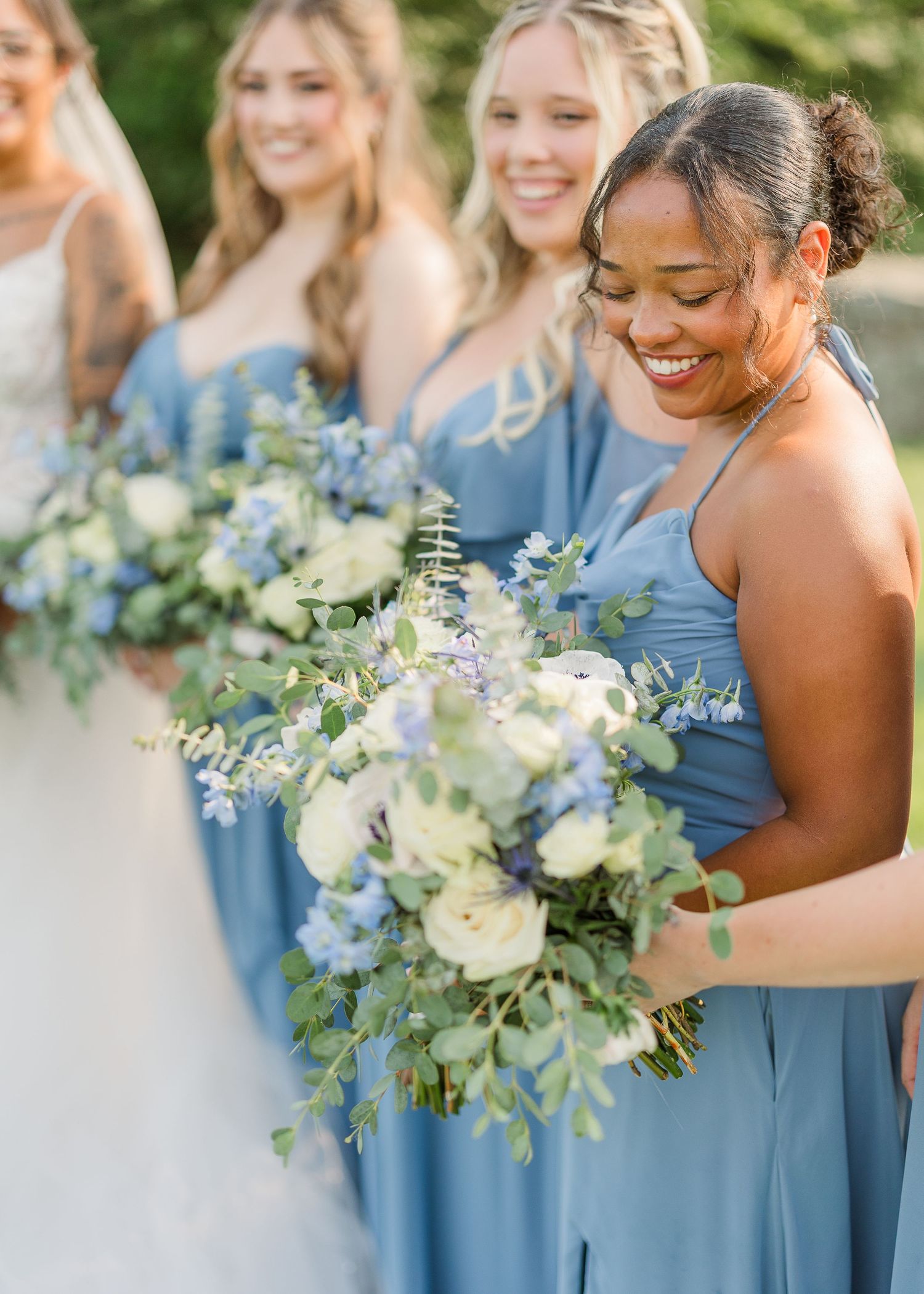 Bridesmaids wearing dusty blue dresses carry elegant white and blue floral bouquets at an outdoor wedding.
