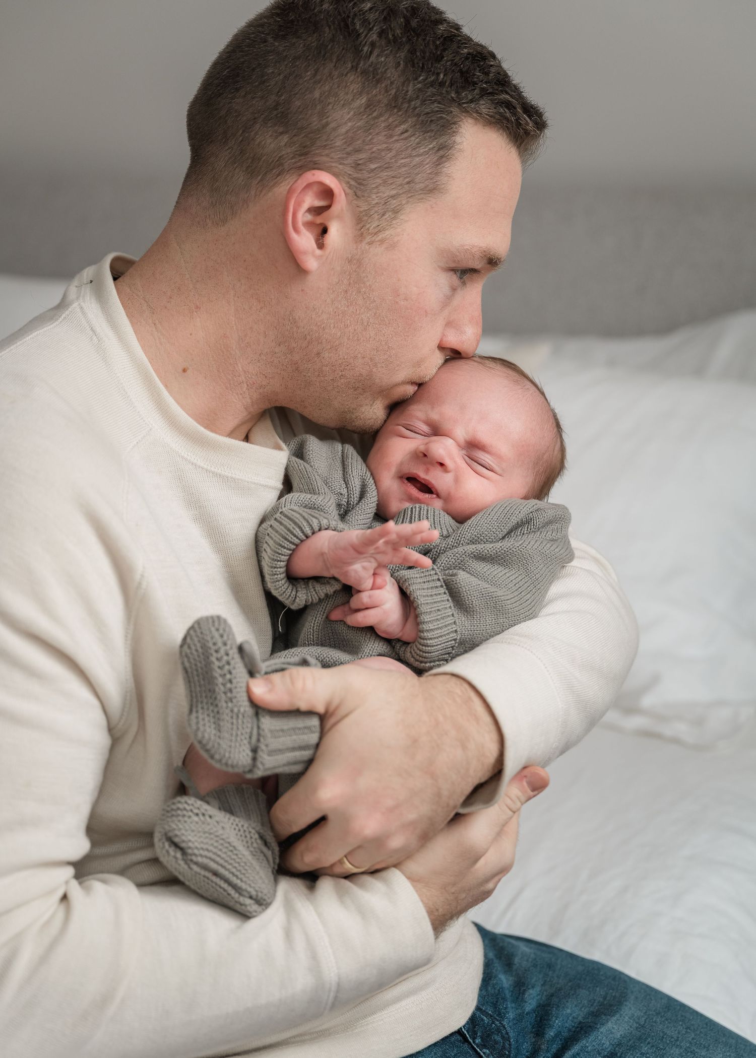 Parent in cream sweater tenderly holding and kissing newborn baby in gray knit sweater on white bedding.