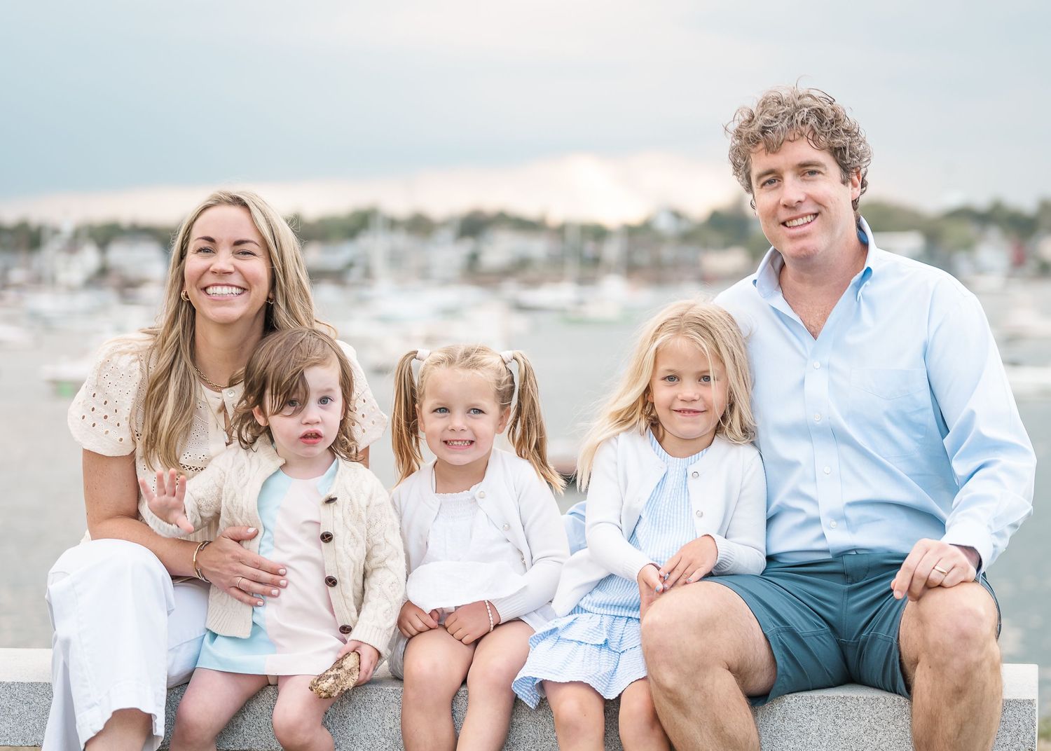 Family wearing coordinated blue and white outfits sits together on a sandy beach at sunset.