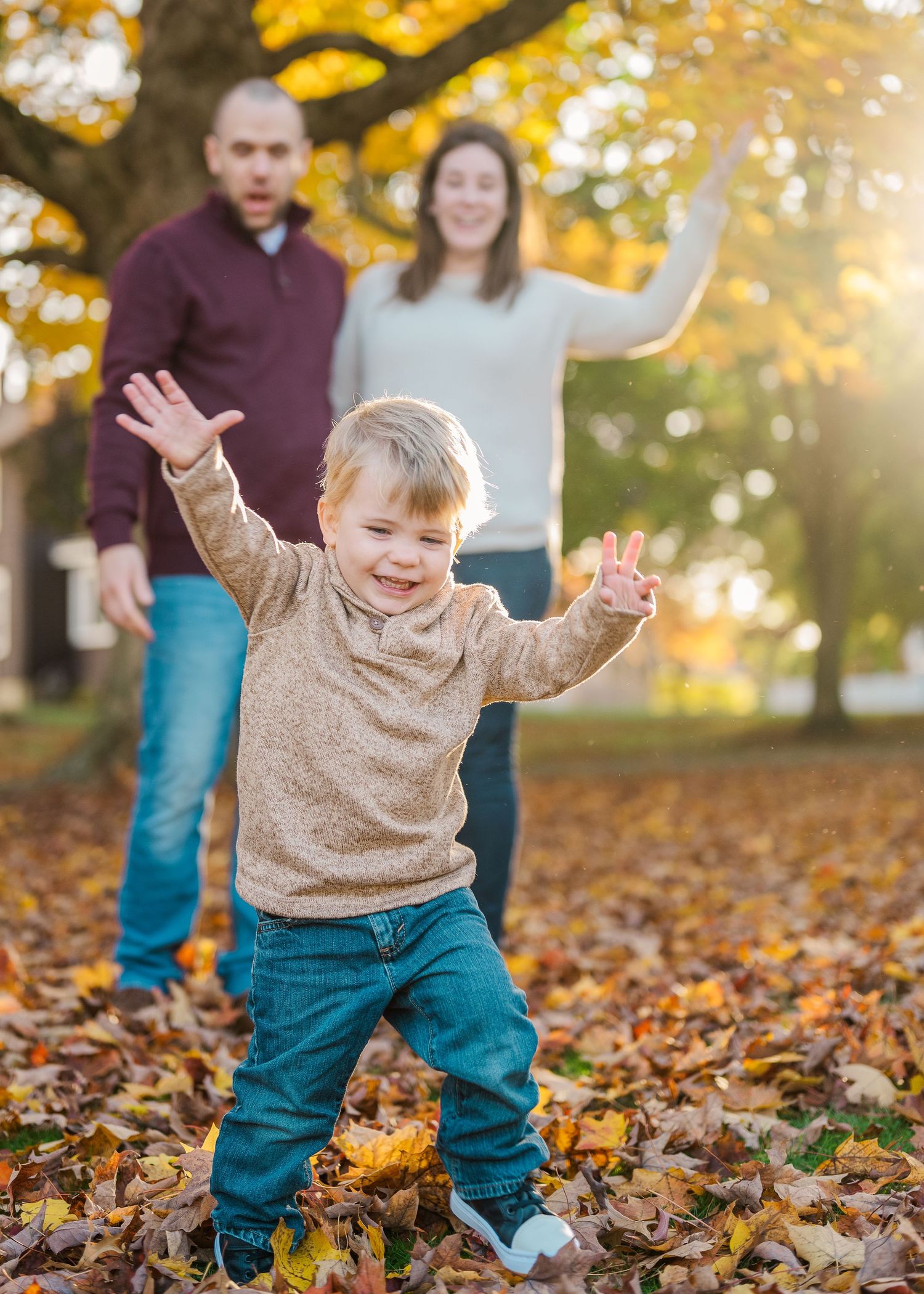 A joyful toddler dances in fallen leaves as adults watch in an autumn park setting.