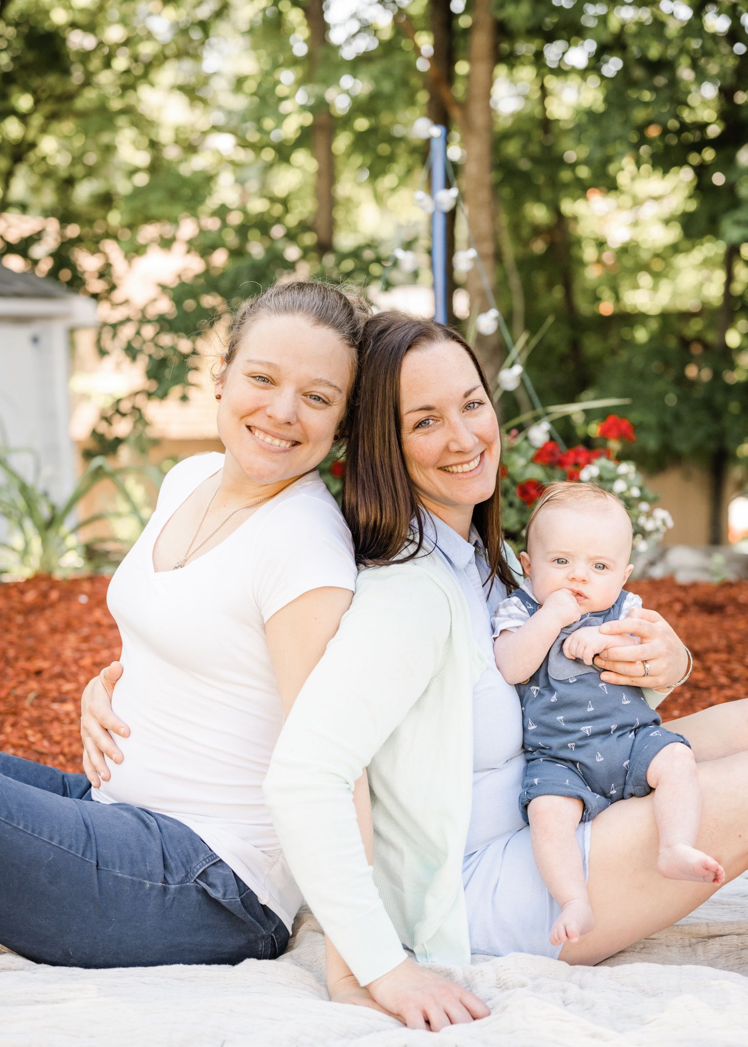 Family sits together on white blanket in garden with red mulch and trees.