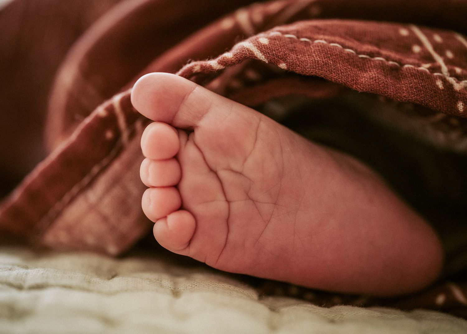 A tiny baby foot peeks out from under a brown blanket in a cozy close-up shot.