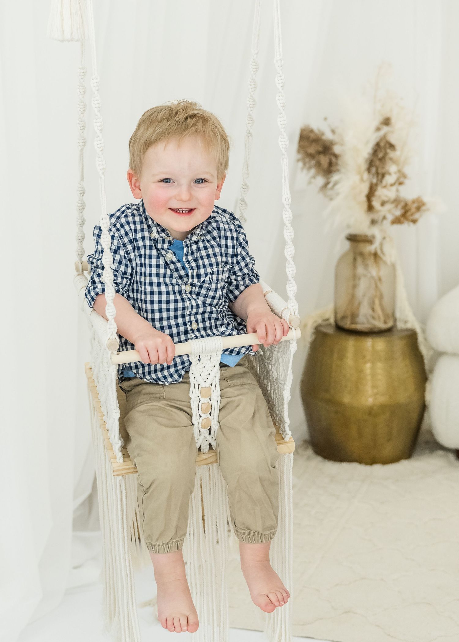Barefoot child wearing gingham shirt and khaki shorts sits on decorative white swing with vase in background.
